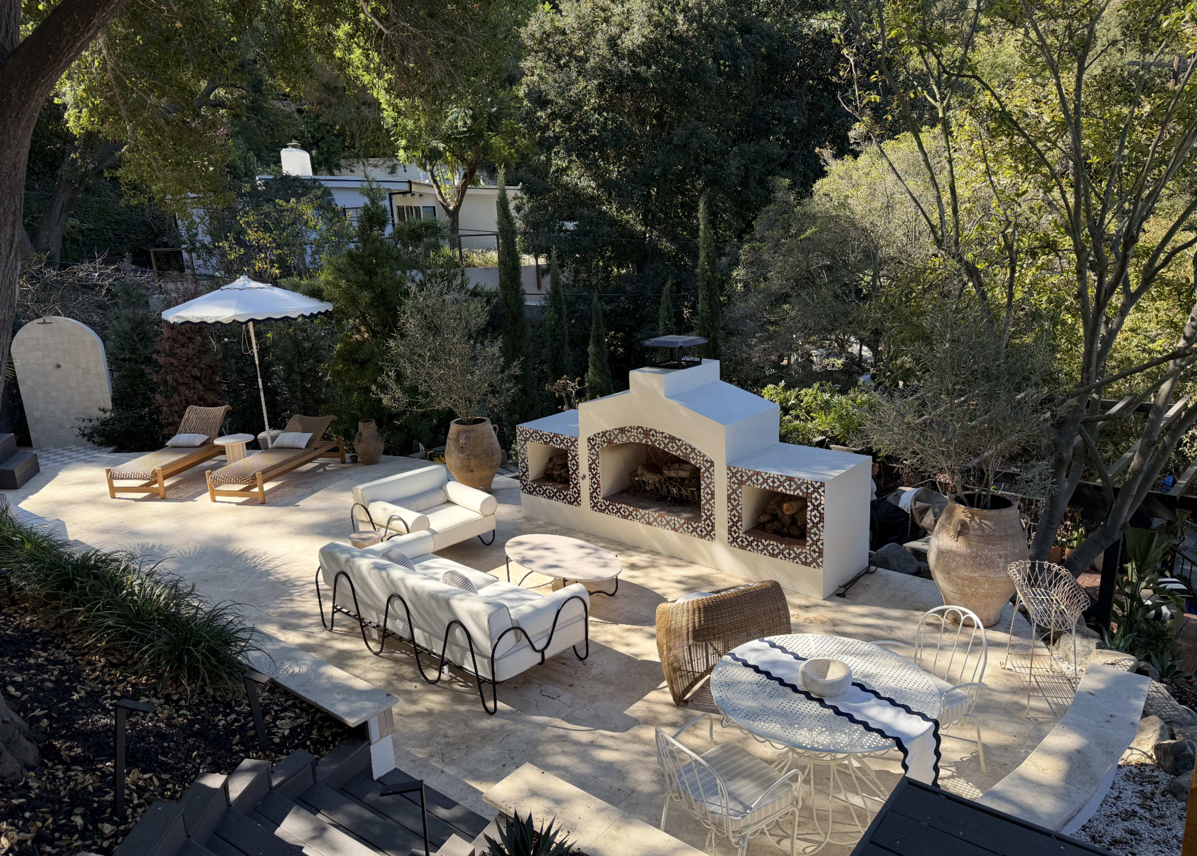 The image shows a landscaped outdoor area with various seating arrangements, including lounge chairs and a white sofa, surrounded by trees and decorative elements like pots and a fireplace.