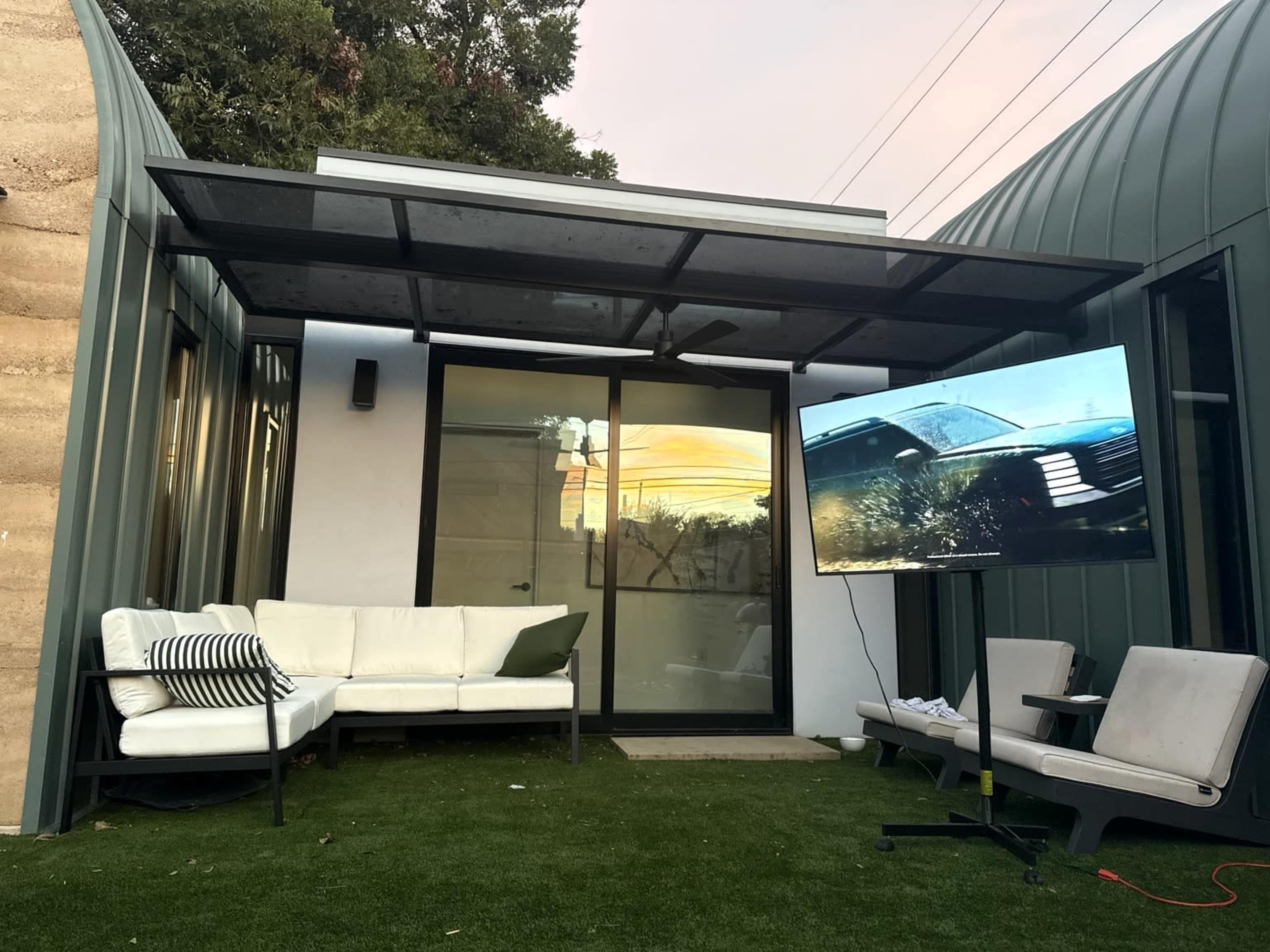 The image shows a modern outdoor seating area with a white couch, a television on a stand, and a green lawn area in front of a house with large glass doors.