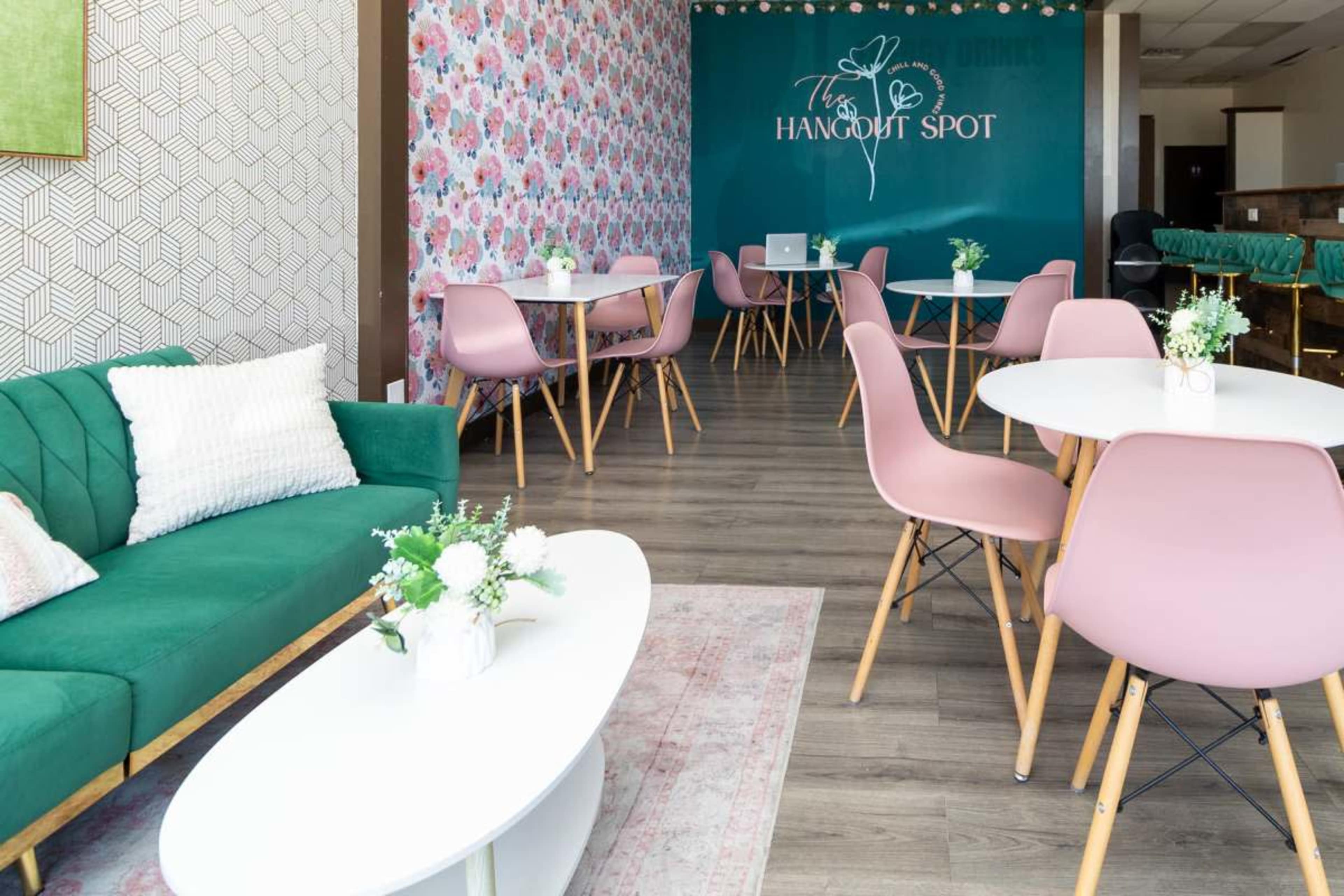 The image shows a modern café interior with green seating, pink chairs, and white tables arranged around a decorative wall featuring blue and floral patterns.