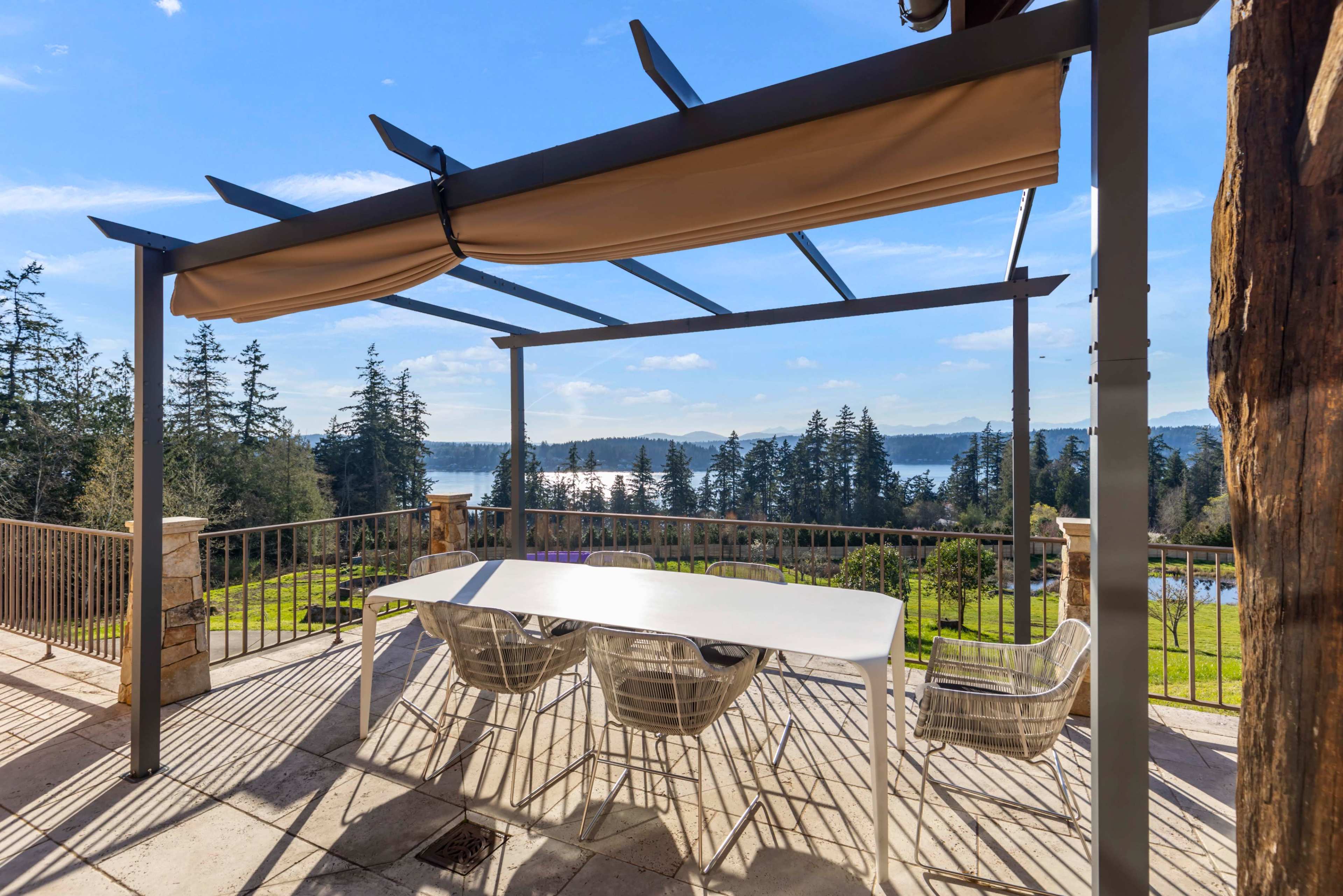 A covered outdoor dining area with a white table and chairs overlooks a lush landscape and water in the distance.