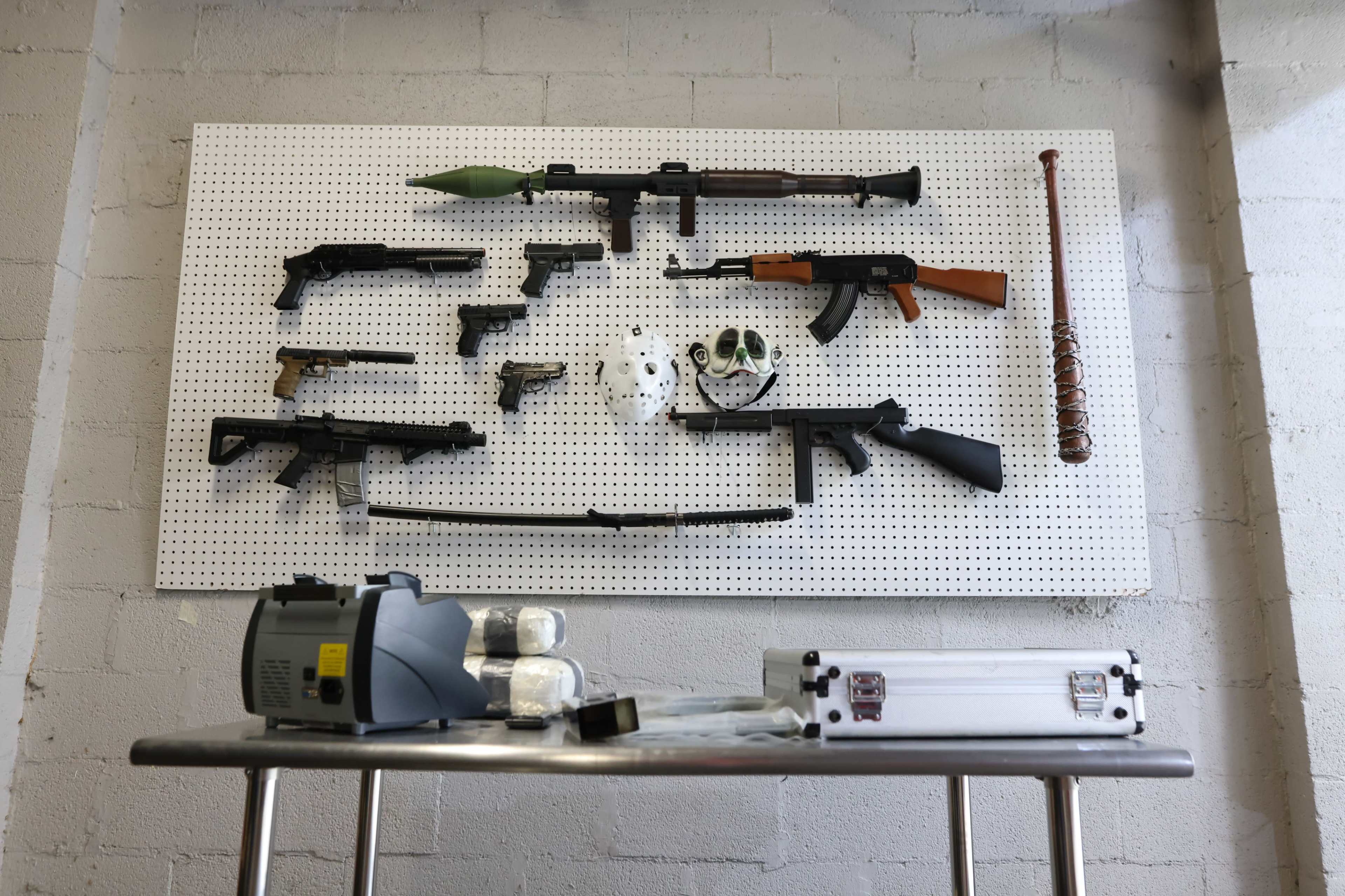 The image shows a display of various firearms and equipment mounted on a pegboard, with some items laid out on a table below.