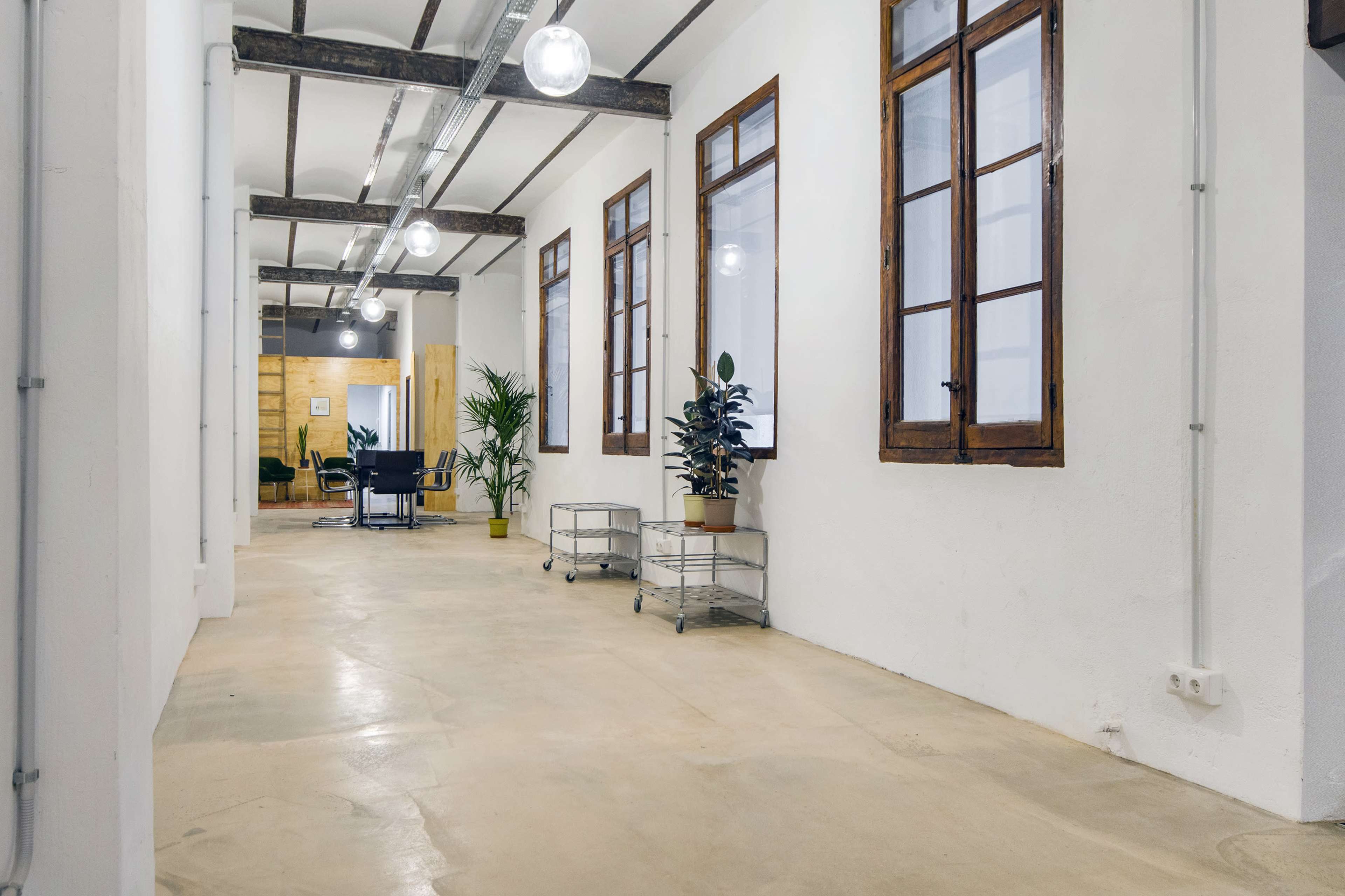 The image shows a well-lit corridor in an office space featuring wooden beams, large windows, and potted plants along the walls.