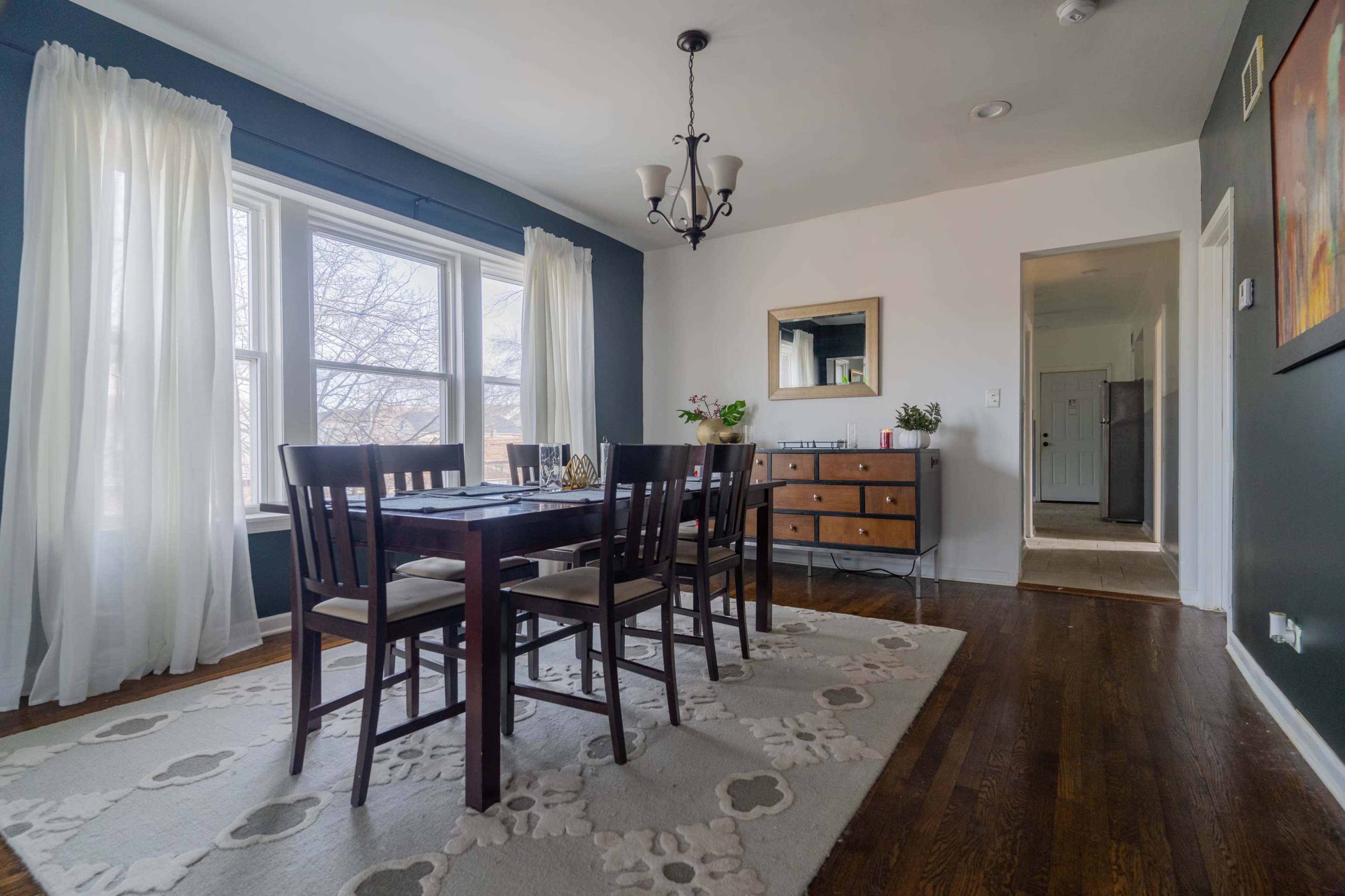 A dining room features a wooden table surrounded by chairs, large windows with sheer curtains, and a sideboard, with an adjoining hallway visible.