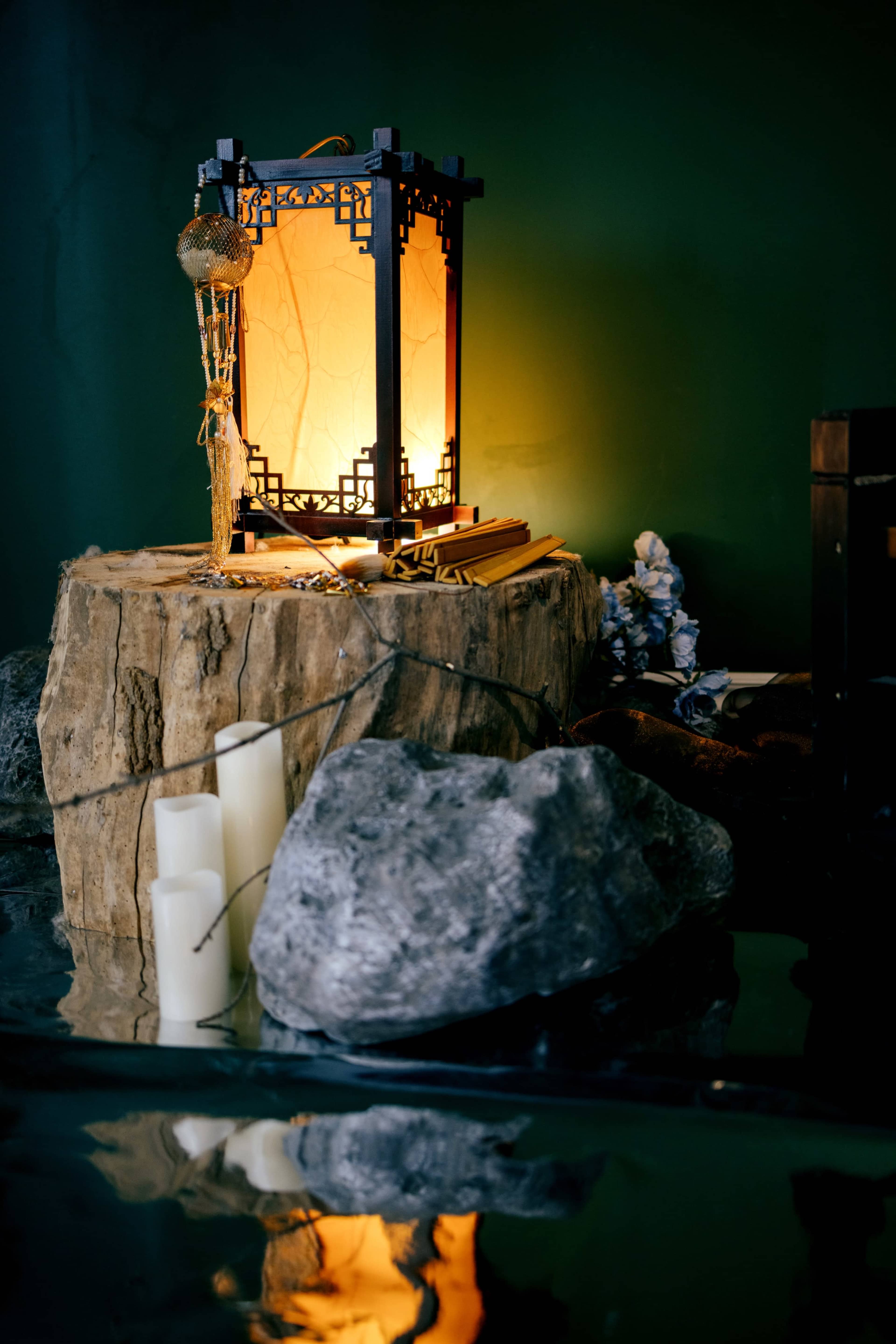 A lantern resting on a wooden stump near several smooth stones and candles arranged around a reflective surface.