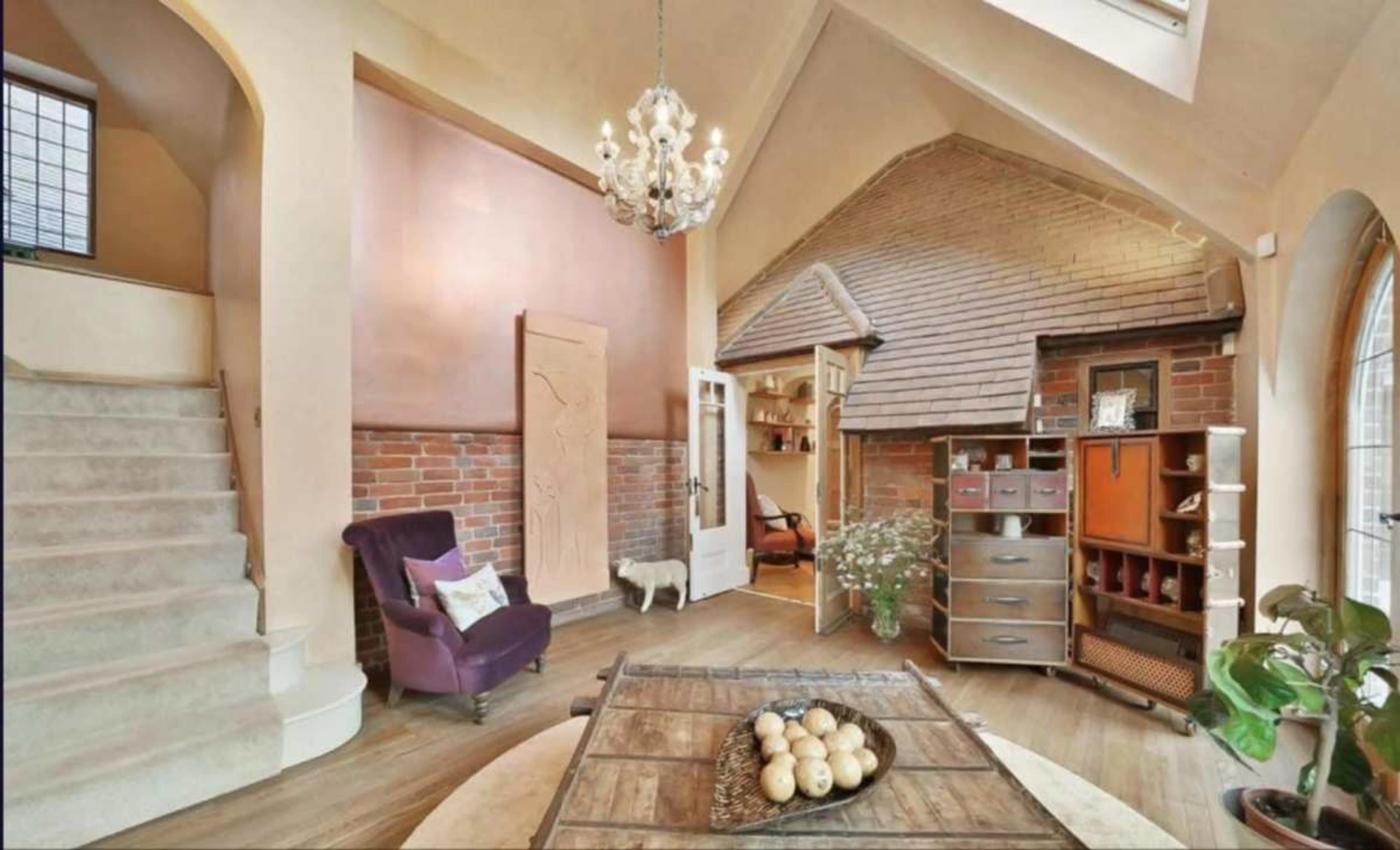 The image shows a cozy entryway with a staircase, a purple armchair, a wooden table with decorative items, and a blend of brick and wooden walls.