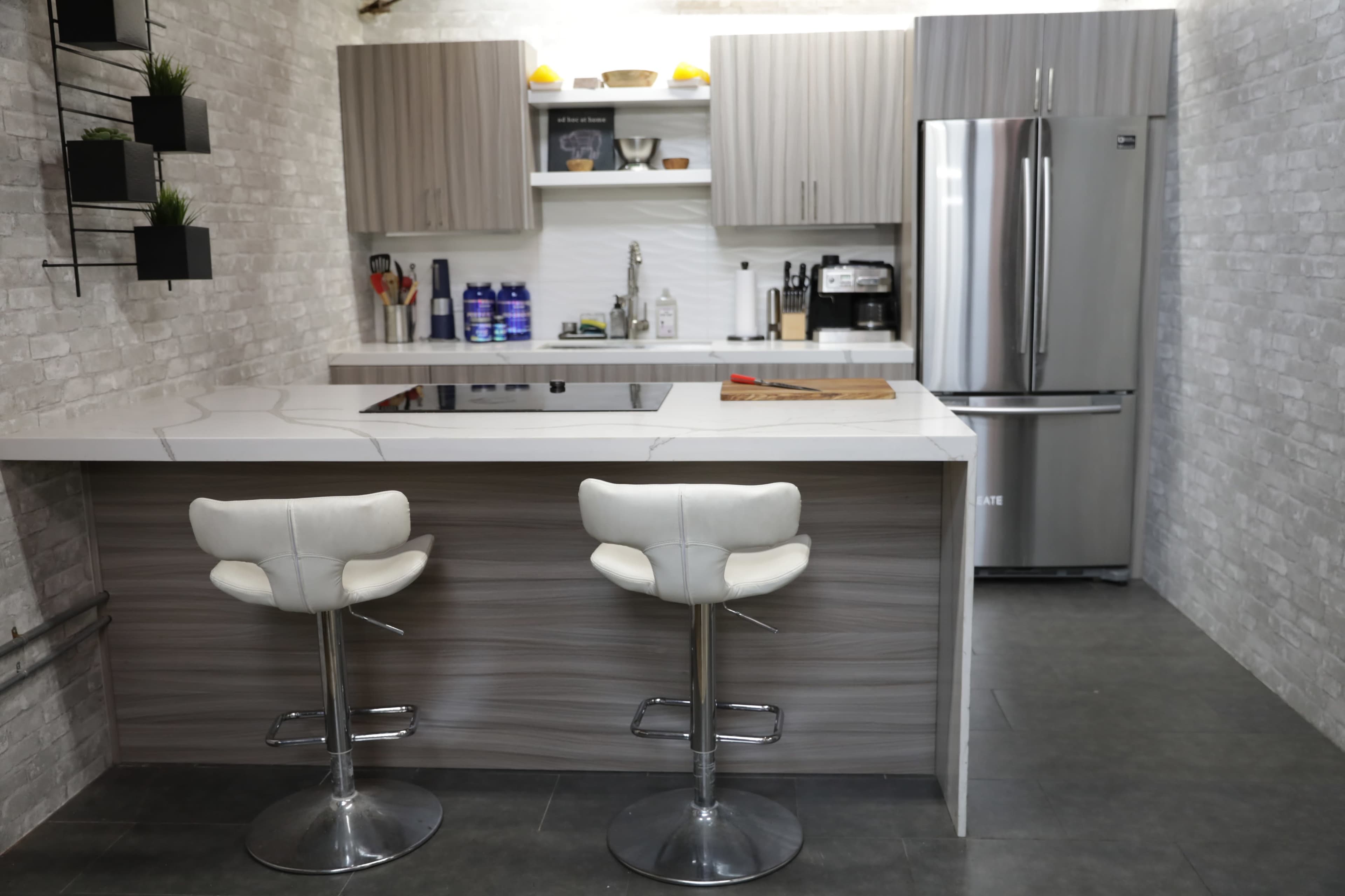 A modern kitchen features gray cabinets, a white countertop with a cooktop, and two white bar stools at a breakfast island.