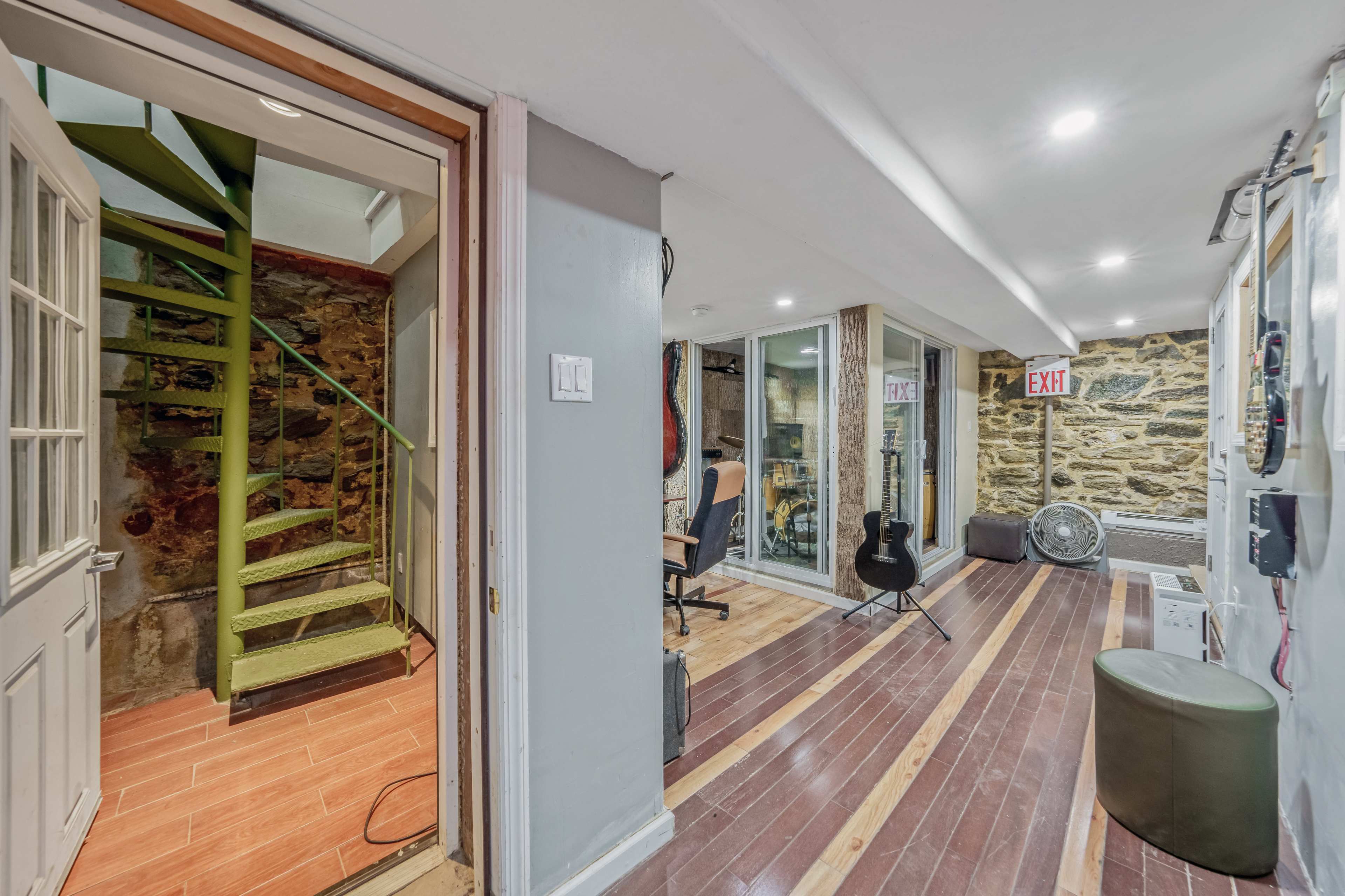 The image shows a basement hallway with exposed stone walls, a staircase, and several functional areas including a workspace and a music setup.