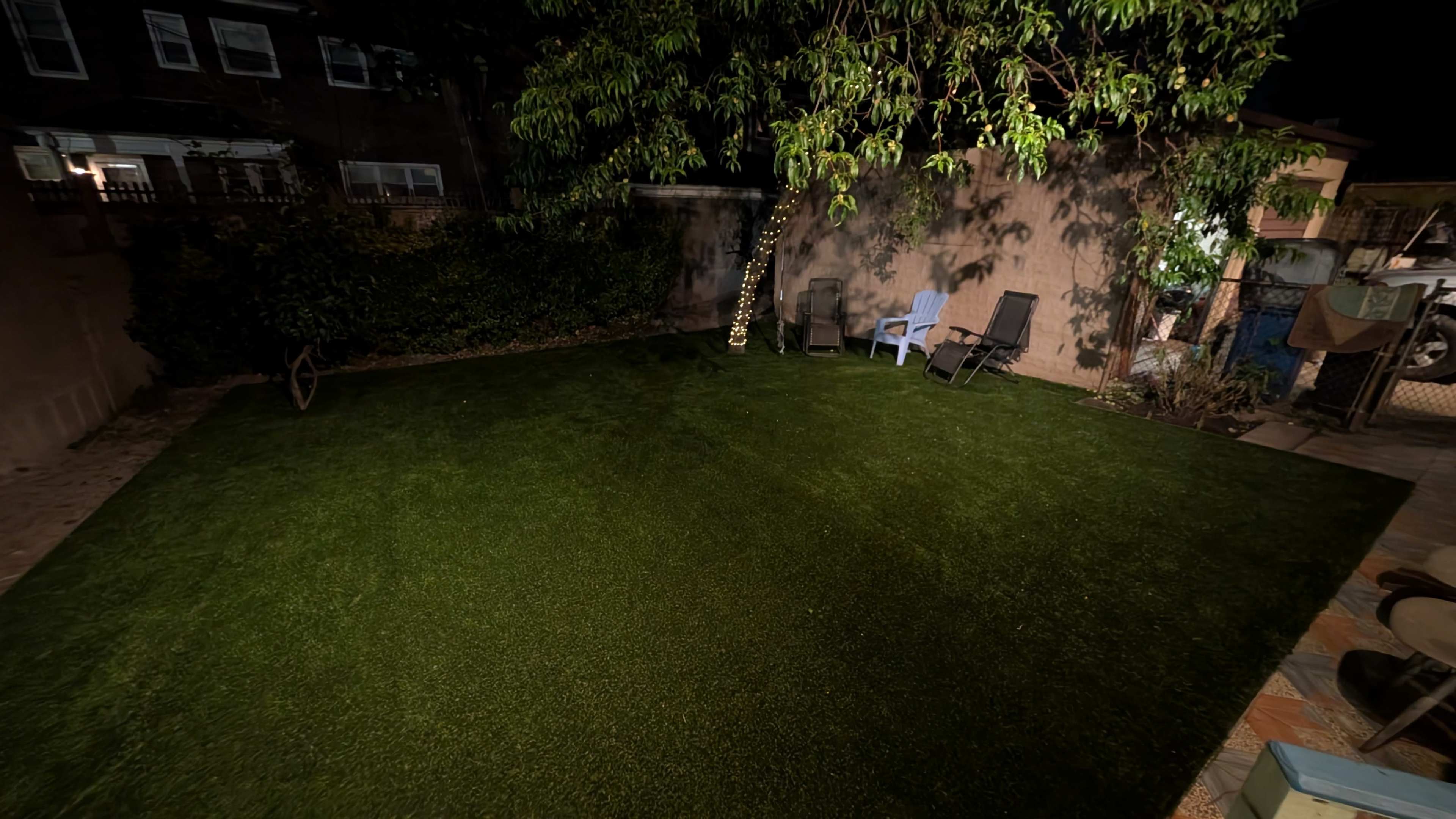 The image shows a well-maintained artificial grass lawn surrounded by a wall and a few chairs under a tree at night.