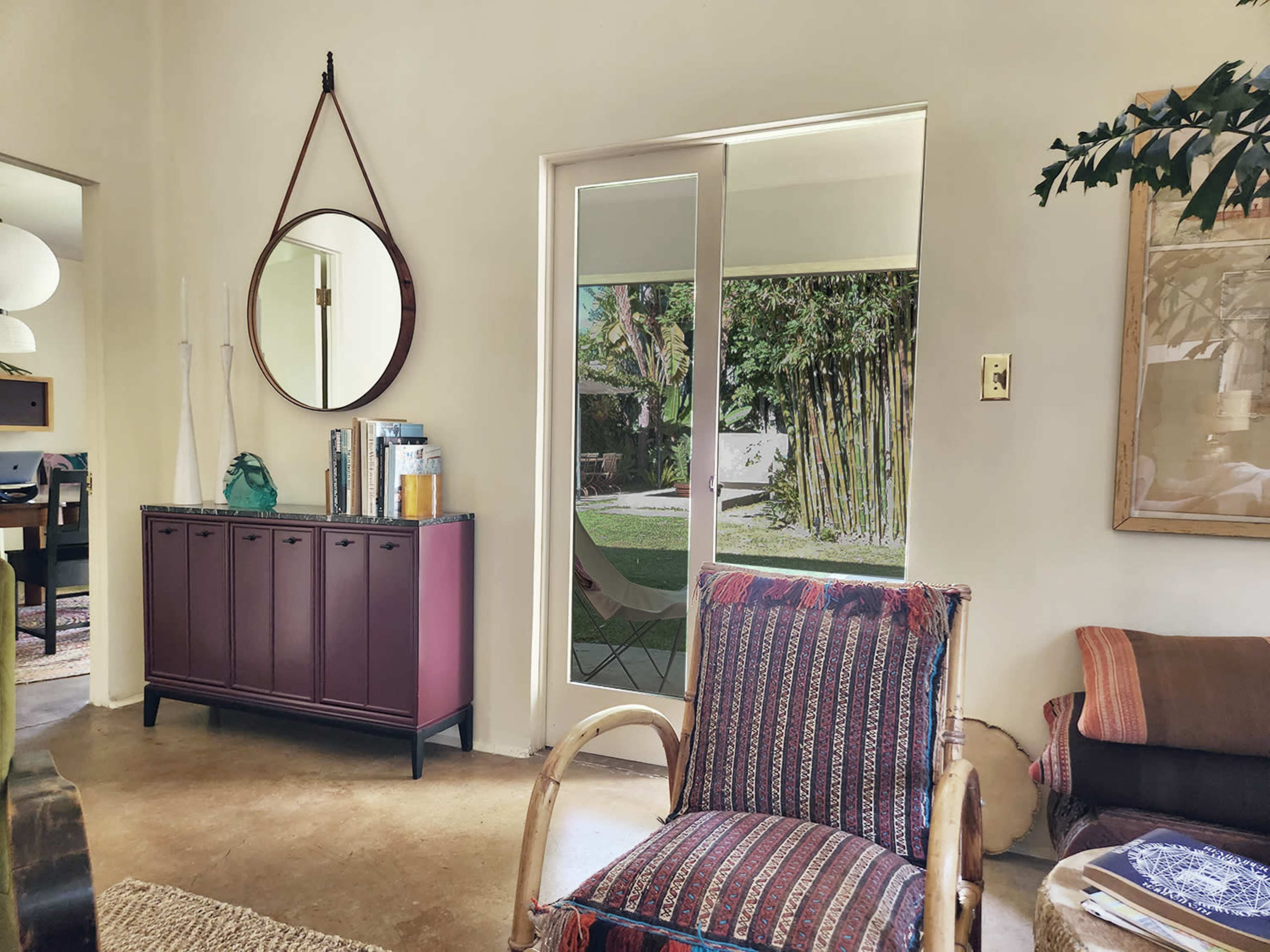A living space featuring a purple cabinet, a round mirror on the wall, and a large glass door leading to a garden area with bamboo.