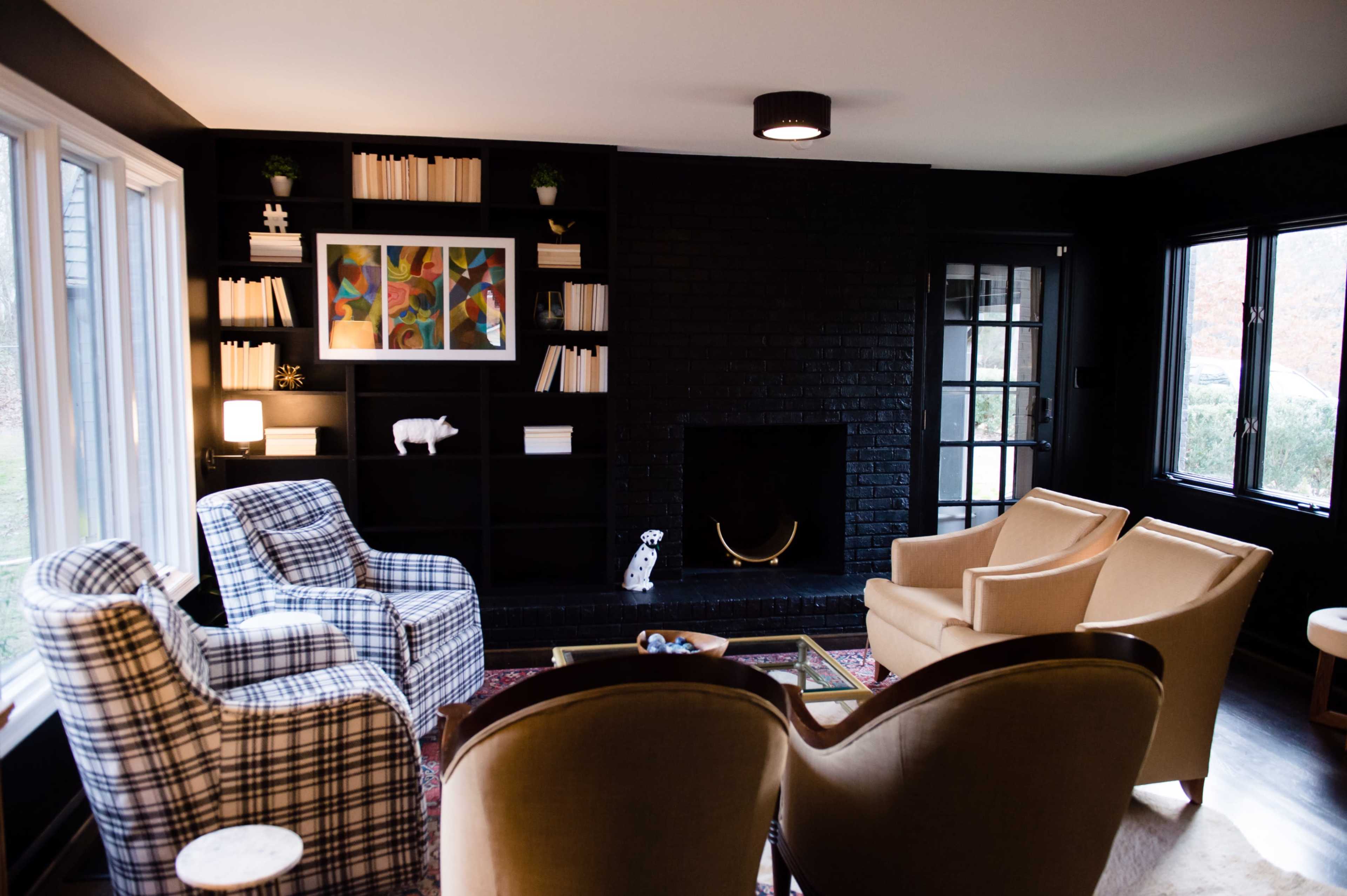 A cozy living room features two patterned armchairs and a beige chair facing a black brick fireplace, with a shelf of books and decorative items in the background.