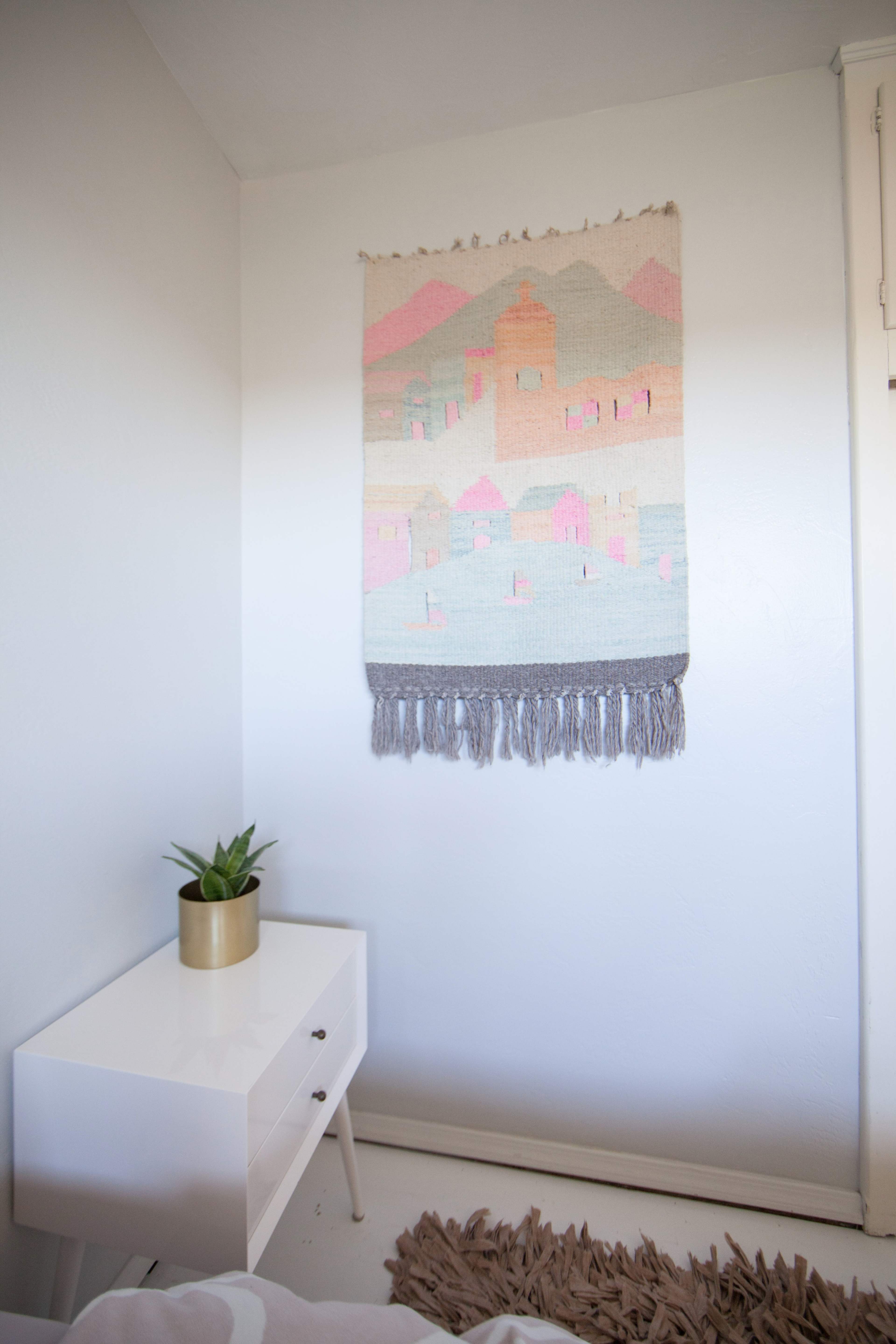 A woven wall tapestry depicting colorful houses hangs above a minimalist white nightstand with a potted plant.