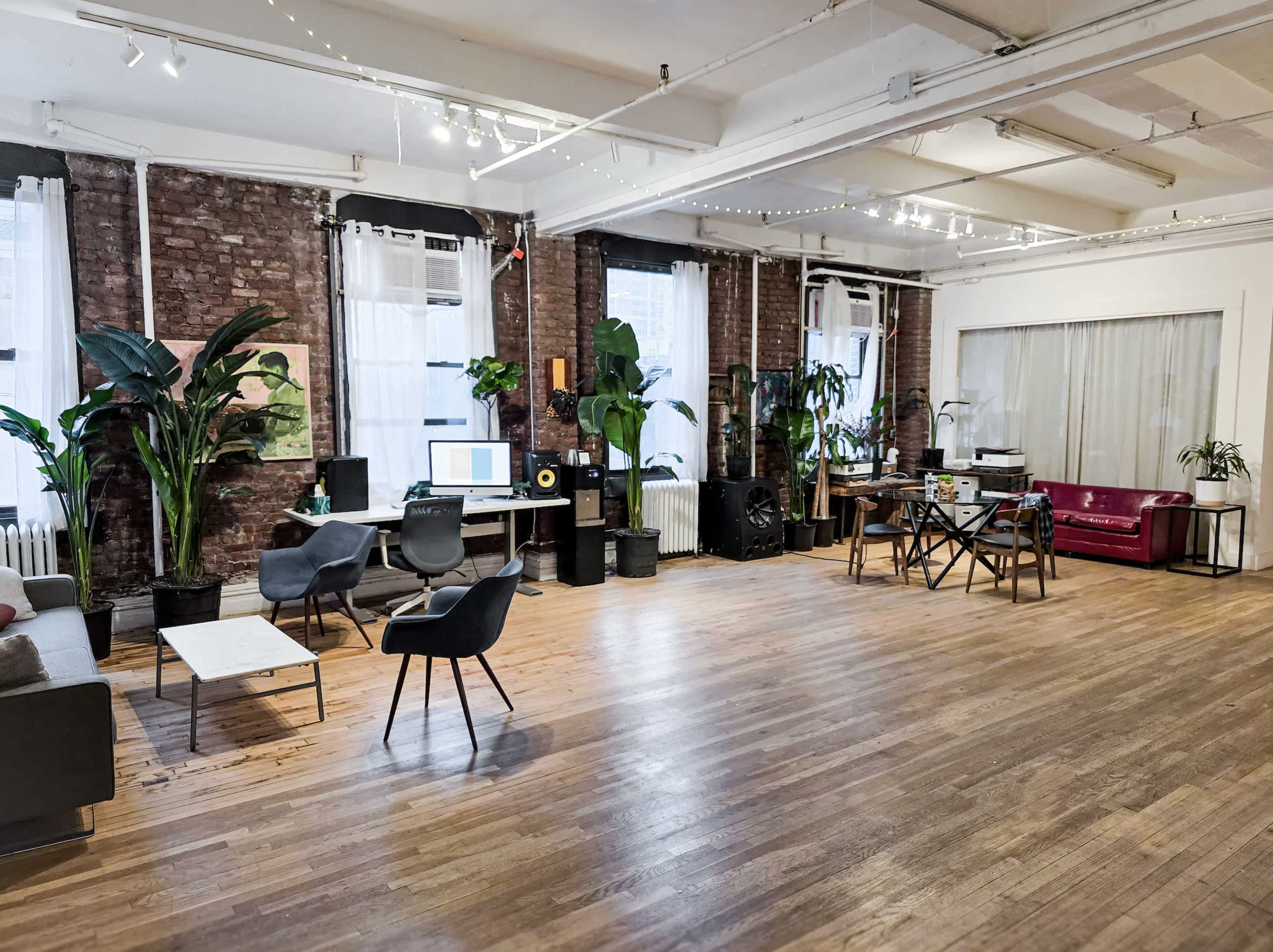 The image shows a spacious, modern studio with wooden flooring, large windows, plants, a combination of seating areas, and workstations.