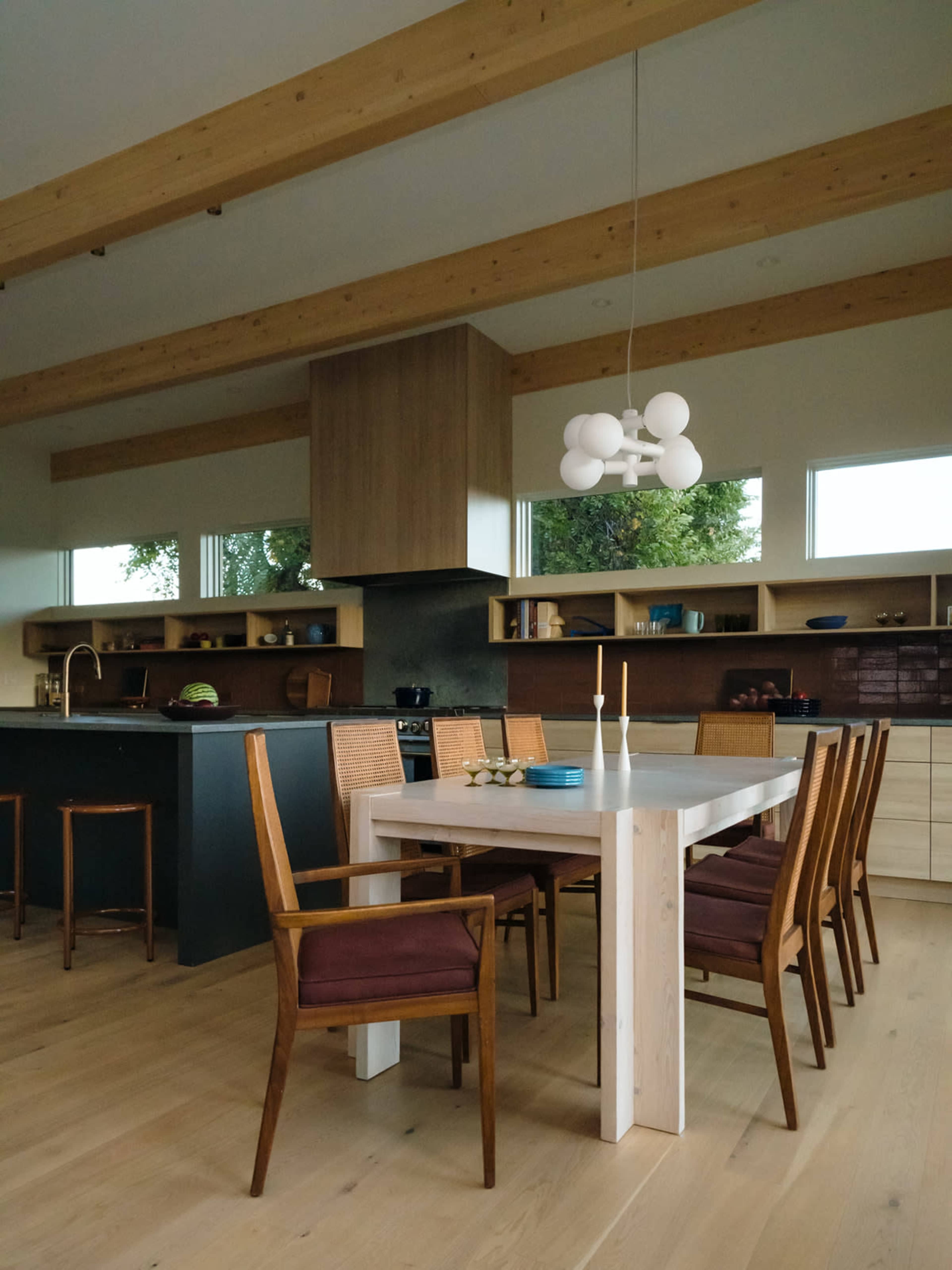 A modern kitchen features a wooden dining table surrounded by eight chairs, with a large hood above a stovetop and ample natural light from the windows.