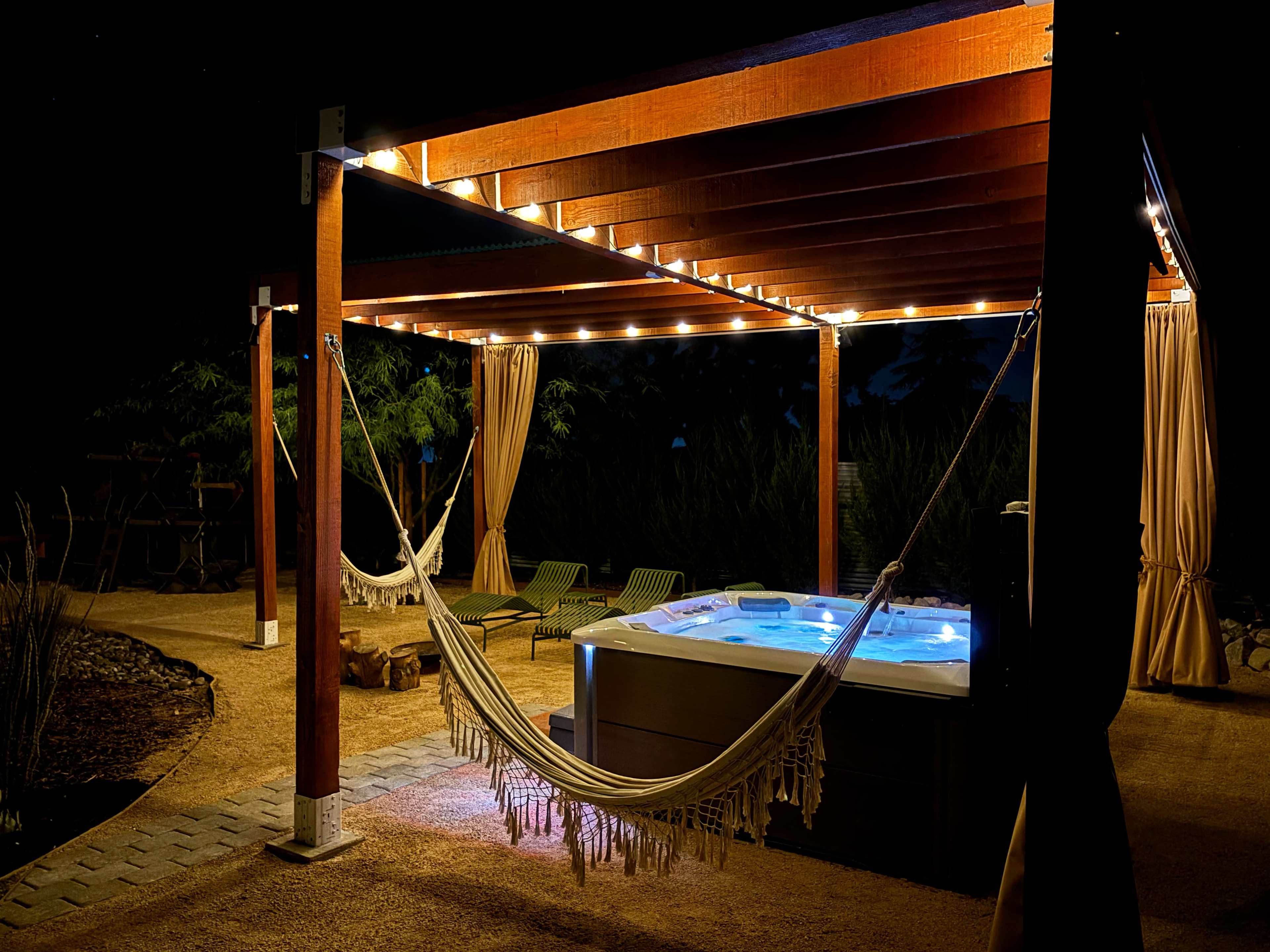 The image shows a hot tub under a wooden pergola illuminated by string lights, with a hammock and lounge chairs nearby.