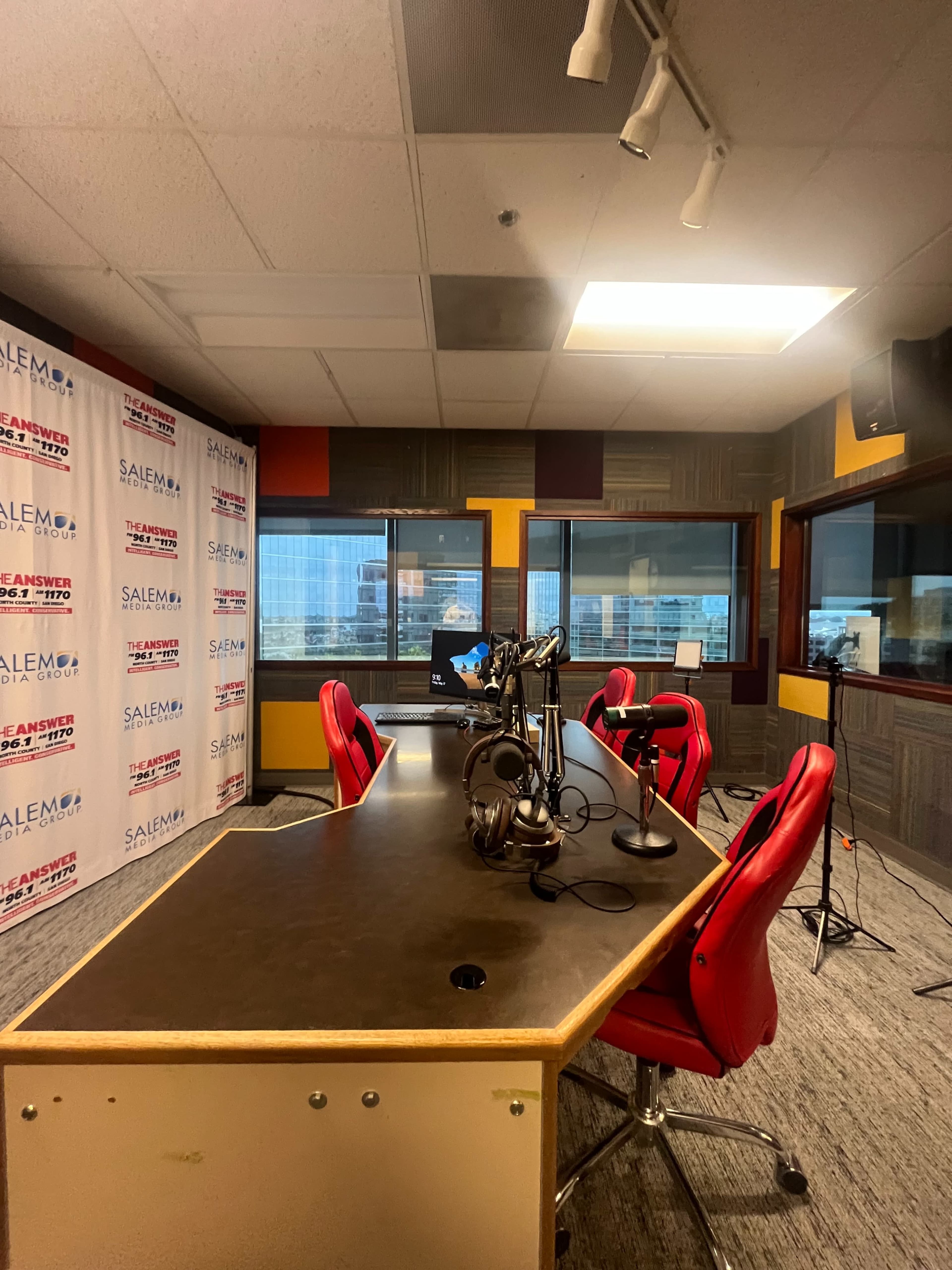 A radio studio with a large table, several red chairs, and microphones, set against a backdrop of windows and branded walls.