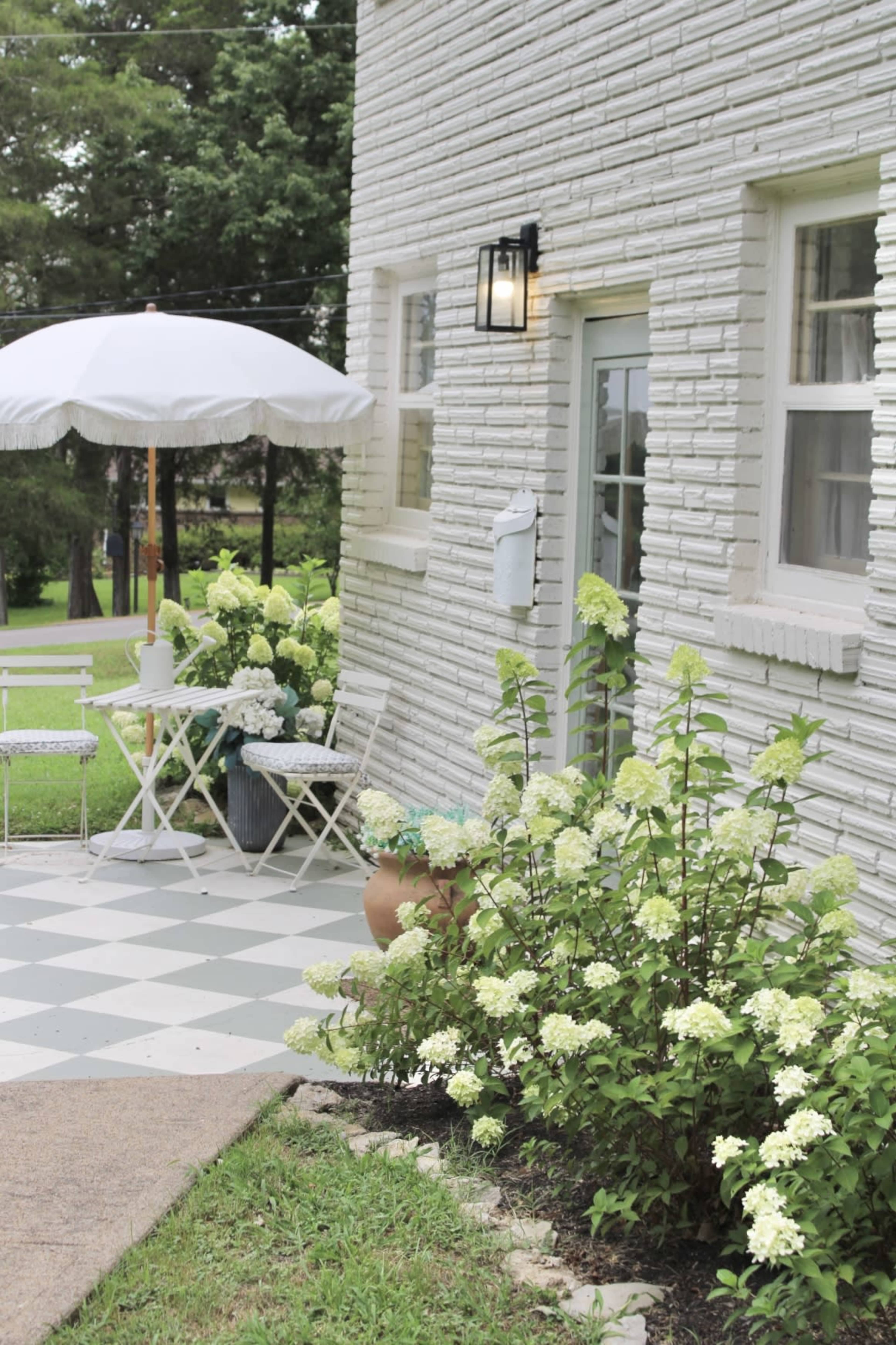 A patio area features a small table and chairs under a white umbrella, with greenery and flowering shrubs alongside a light-colored brick wall.