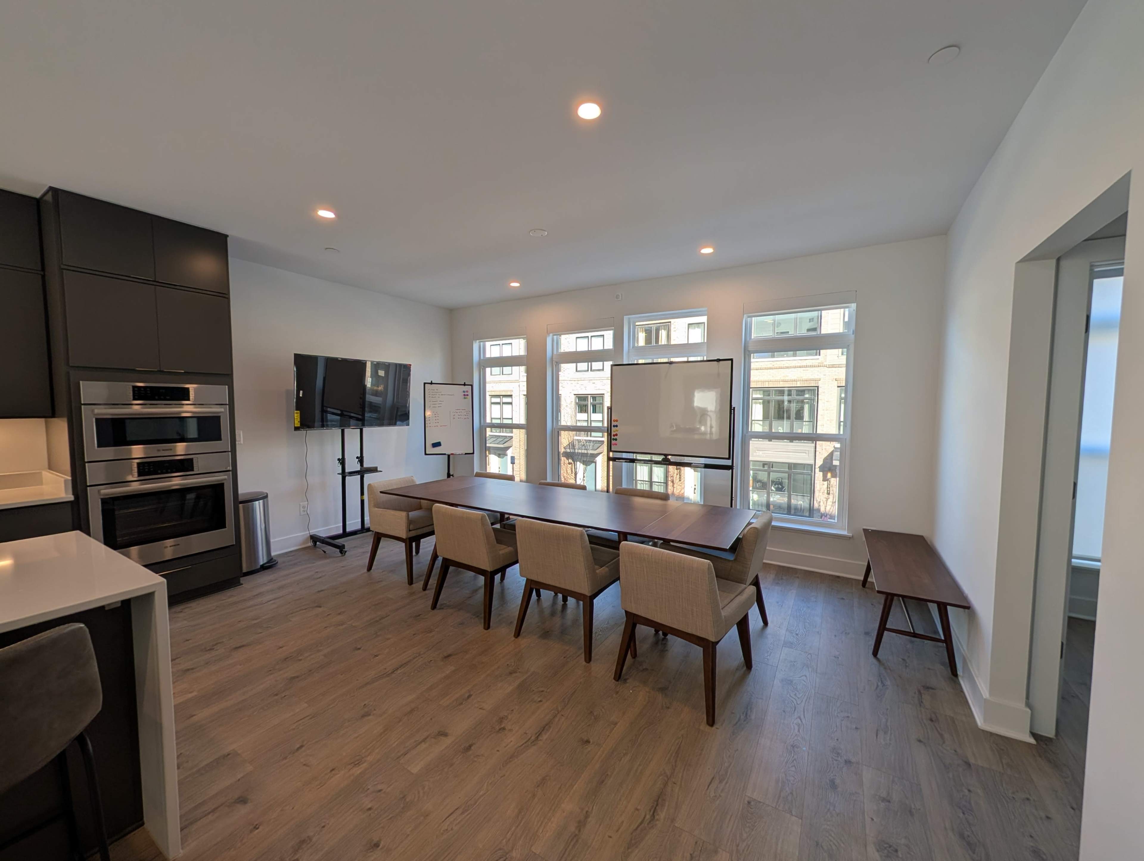 The image shows a modern conference room with a large table, several chairs, a wall-mounted TV, and large windows providing natural light.