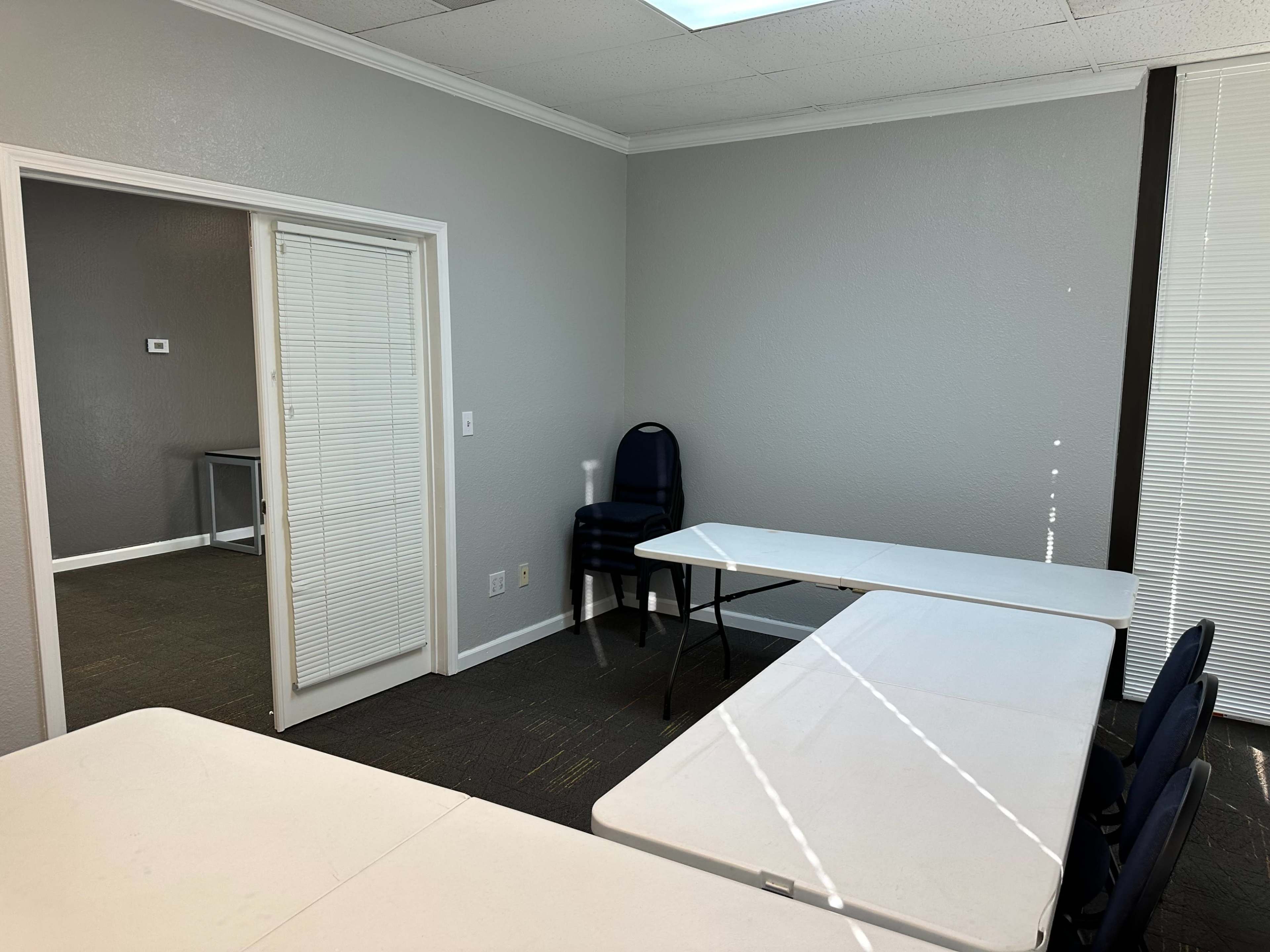 A small meeting room with two white tables arranged in an L-shape, a blue chair stacked against the wall, and a door leading to another room.