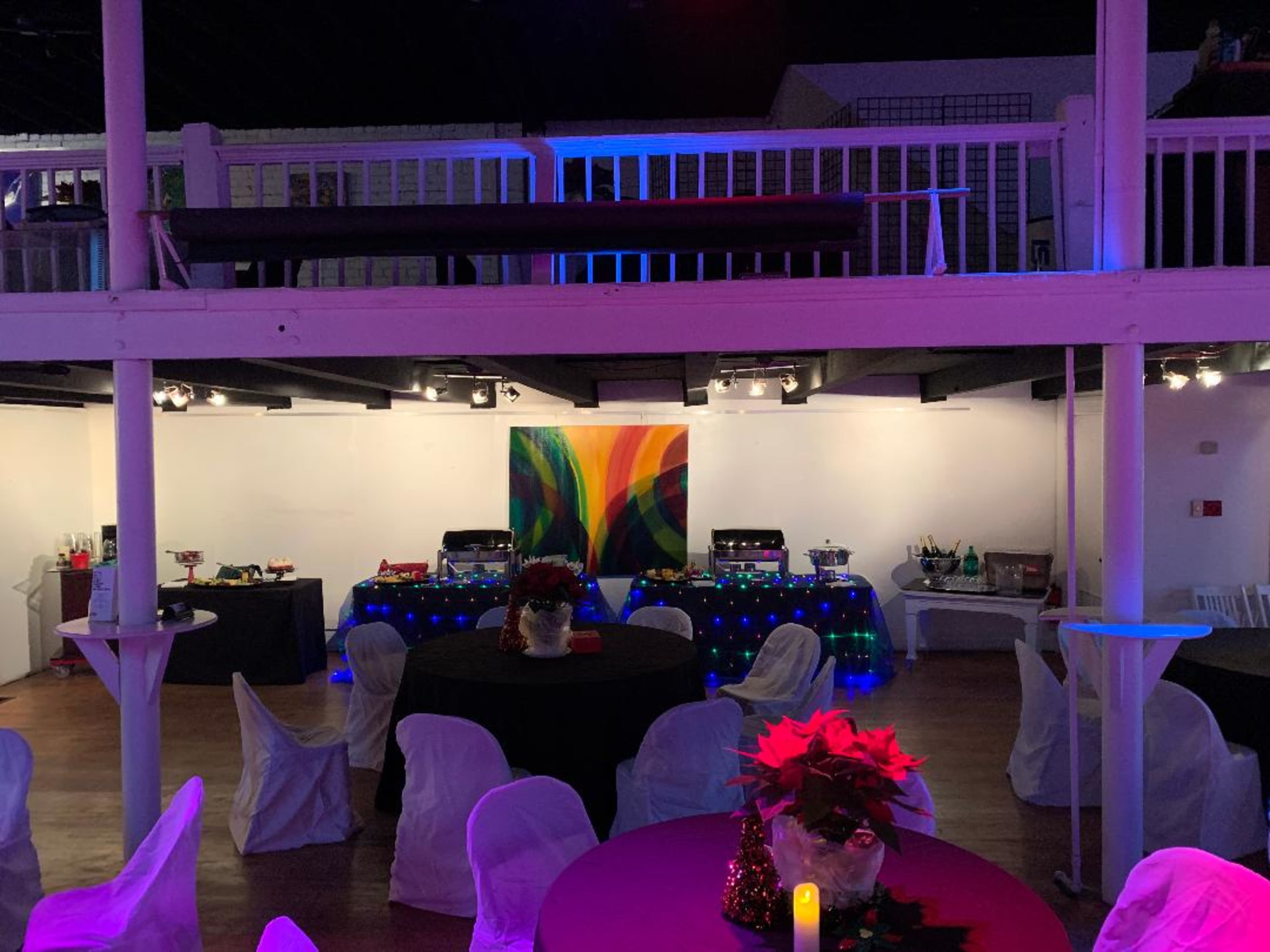 A banquet room is set up with tables adorned in black and purple tablecloths, featuring a colorful abstract painting on the wall and a buffet table filled with food.