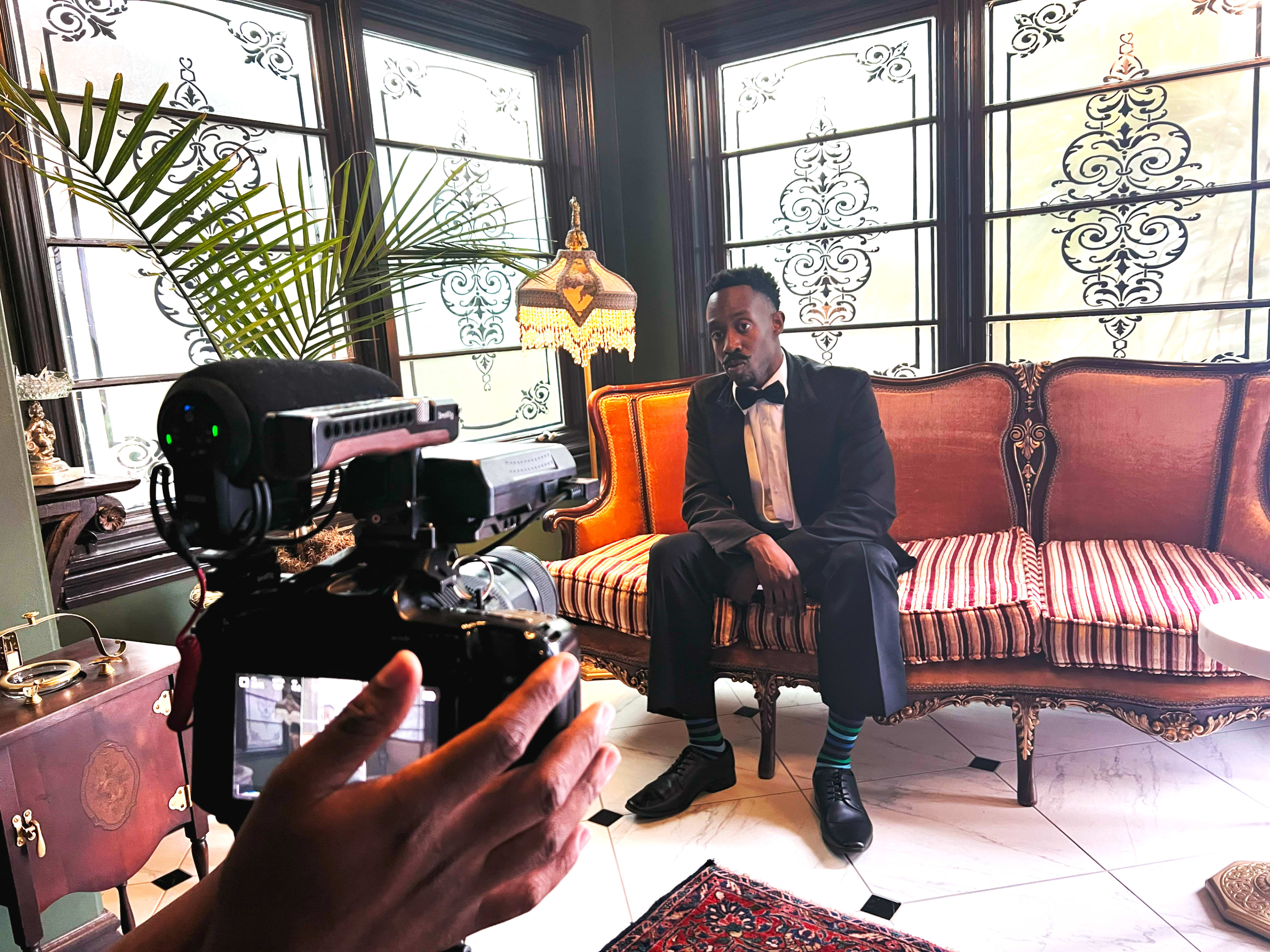 A person dressed in formal attire sits on a striped sofa while a camera is directed at them in a well-decorated room.