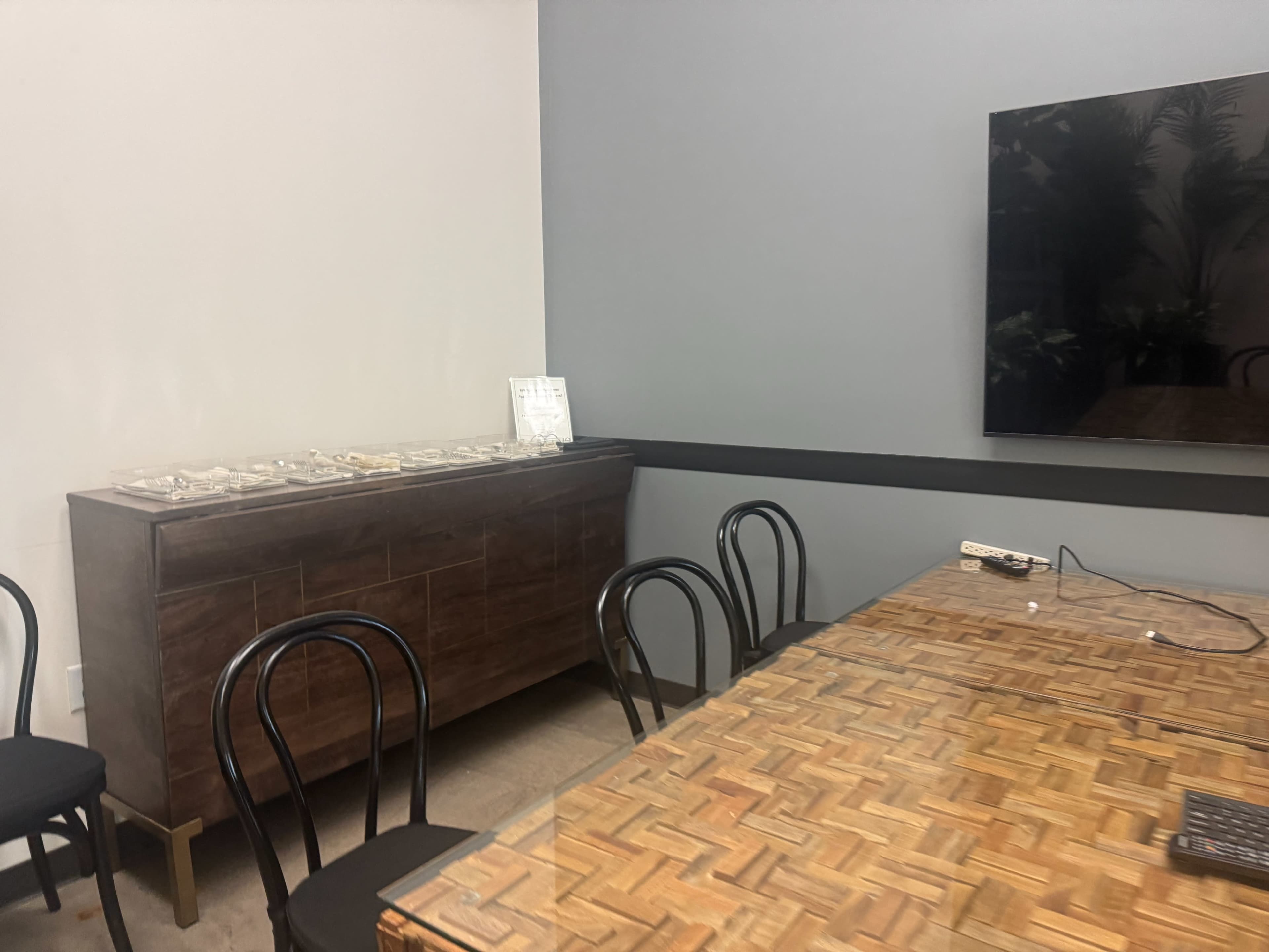 The image shows a meeting room with a wooden table, several black chairs, a cabinet against the wall, and a large television mounted on the wall.