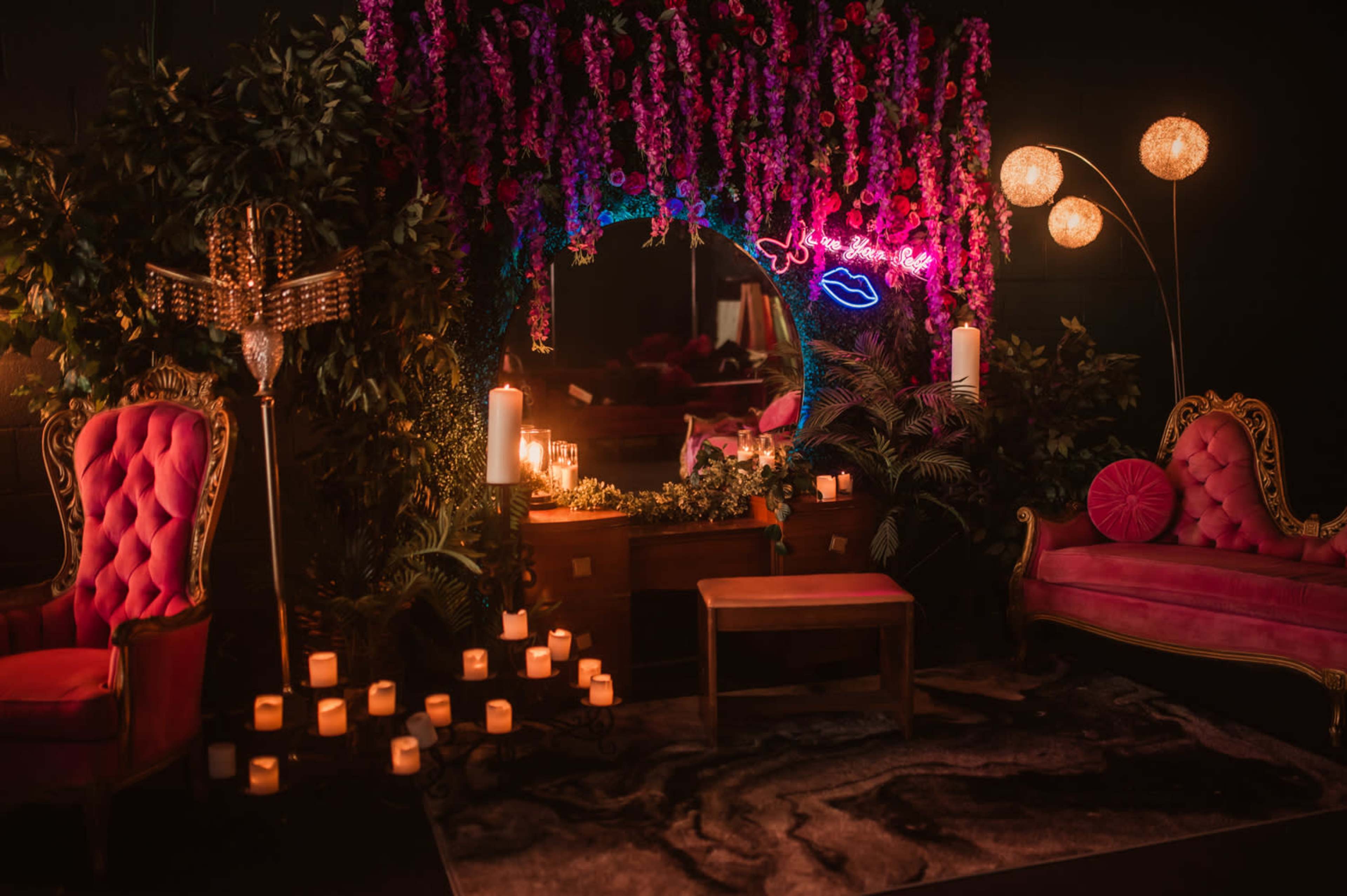 The scene features an elegantly decorated space with pink upholstered furniture, vibrant floral arrangements, candles, and soft lighting.
