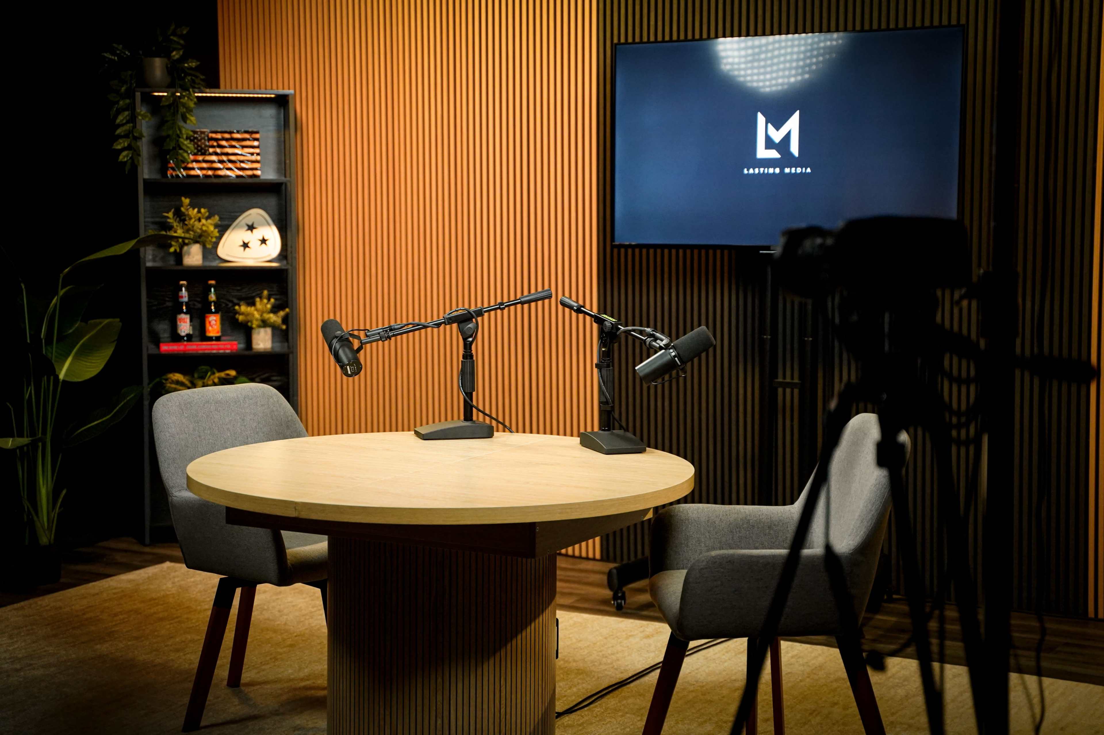 The image shows a minimalistic podcast studio with two microphones positioned on a round wooden table and a screen displaying the "Latidon Media" logo in the background.