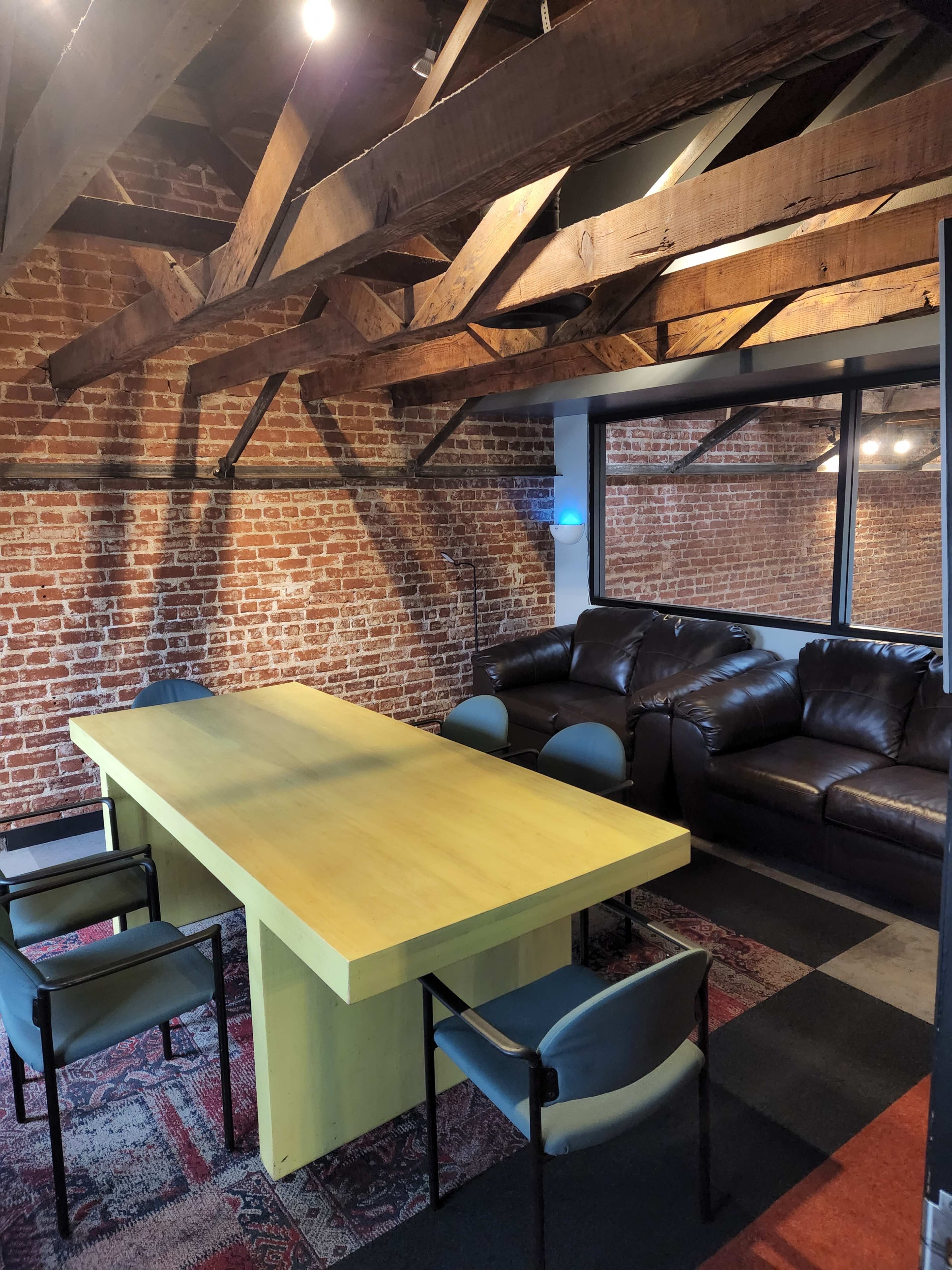 A room with exposed brick walls, a large yellow table surrounded by chairs, and a brown leather sofa along one side.