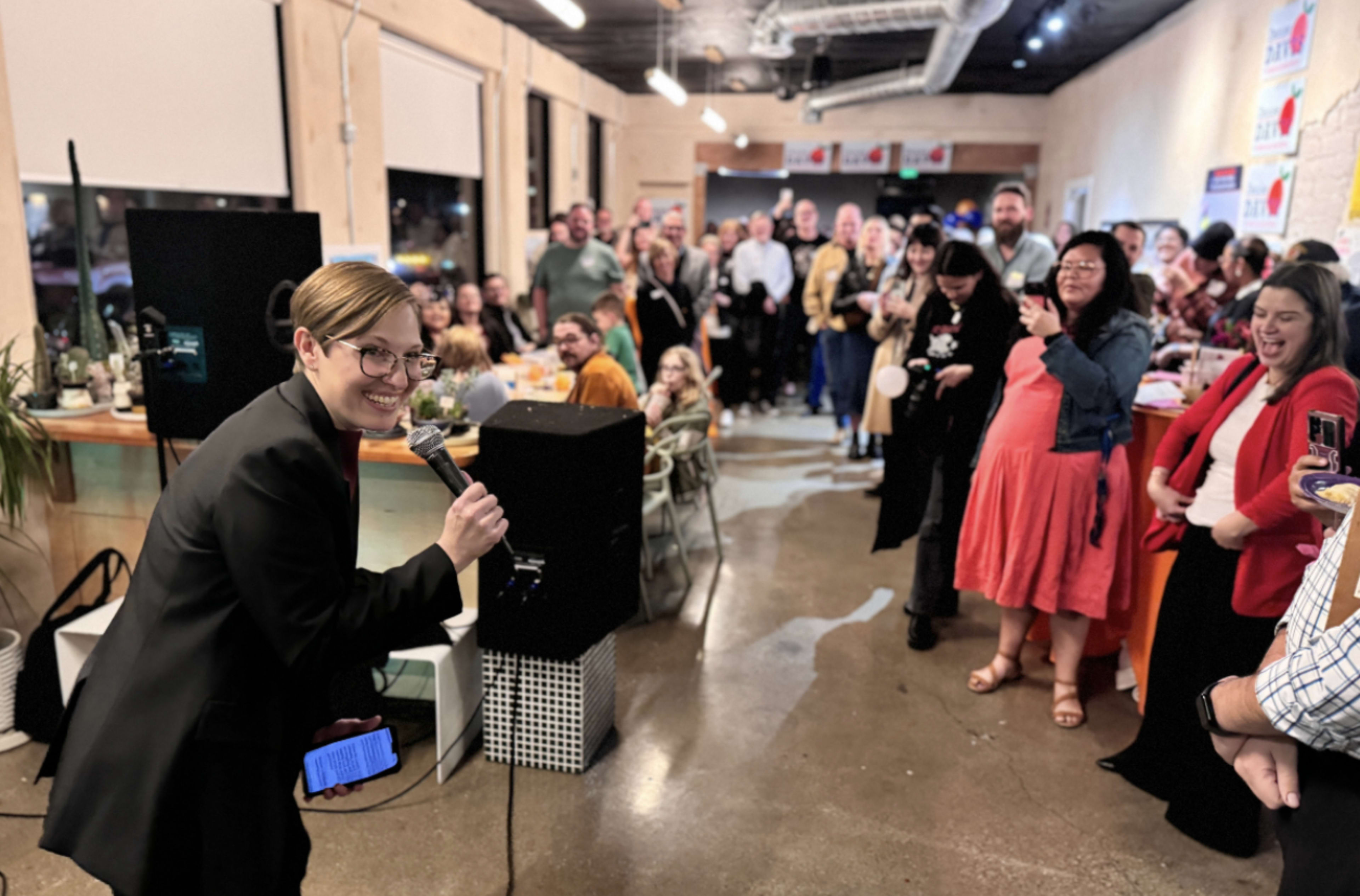 A woman in a black suit holds a microphone and smiles at a gathered crowd during a celebration in a well-lit venue.