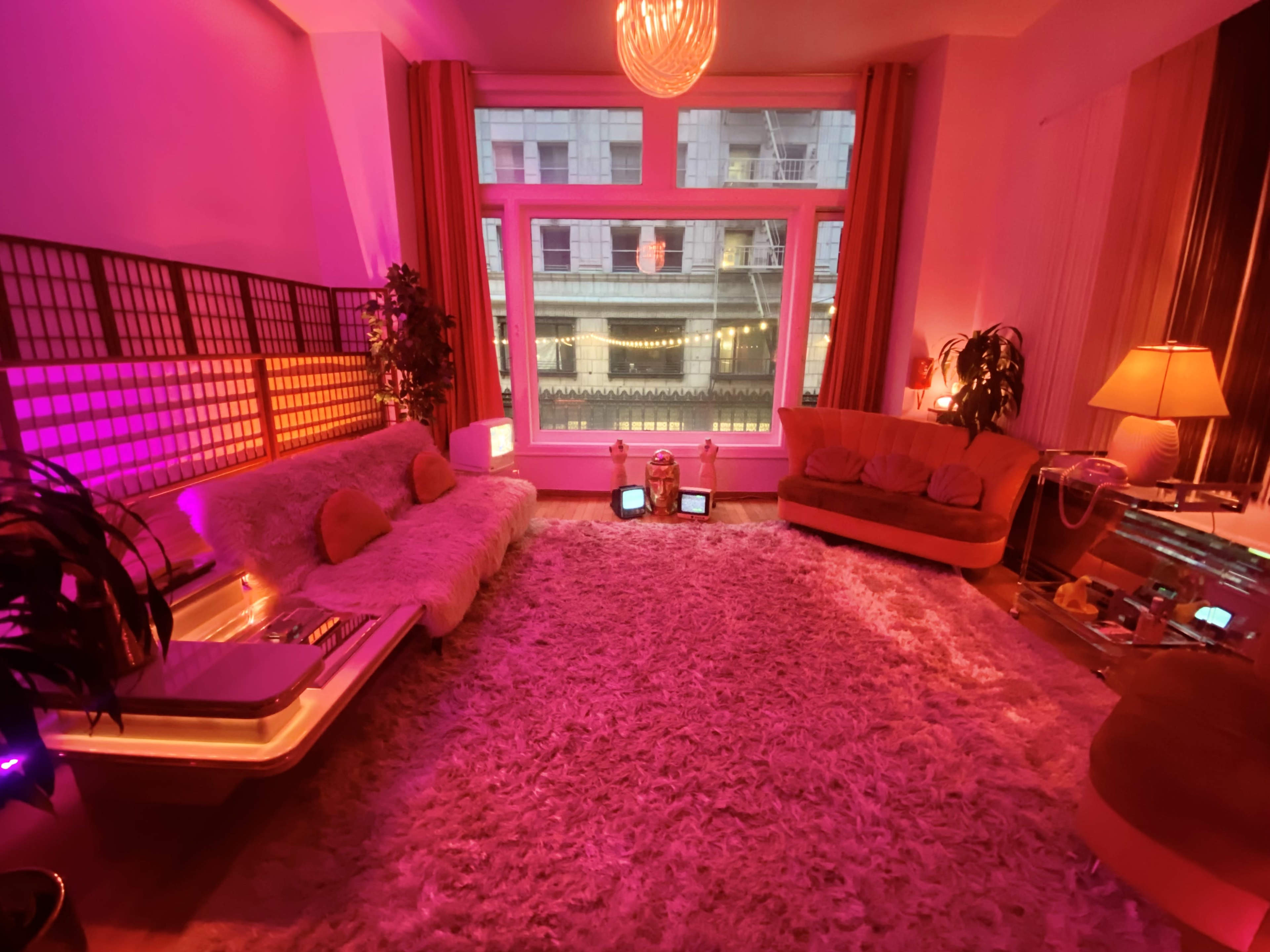 A retro-styled living room with plush furniture, a large area rug, and colorful lighting, featuring a view of a street through large windows.