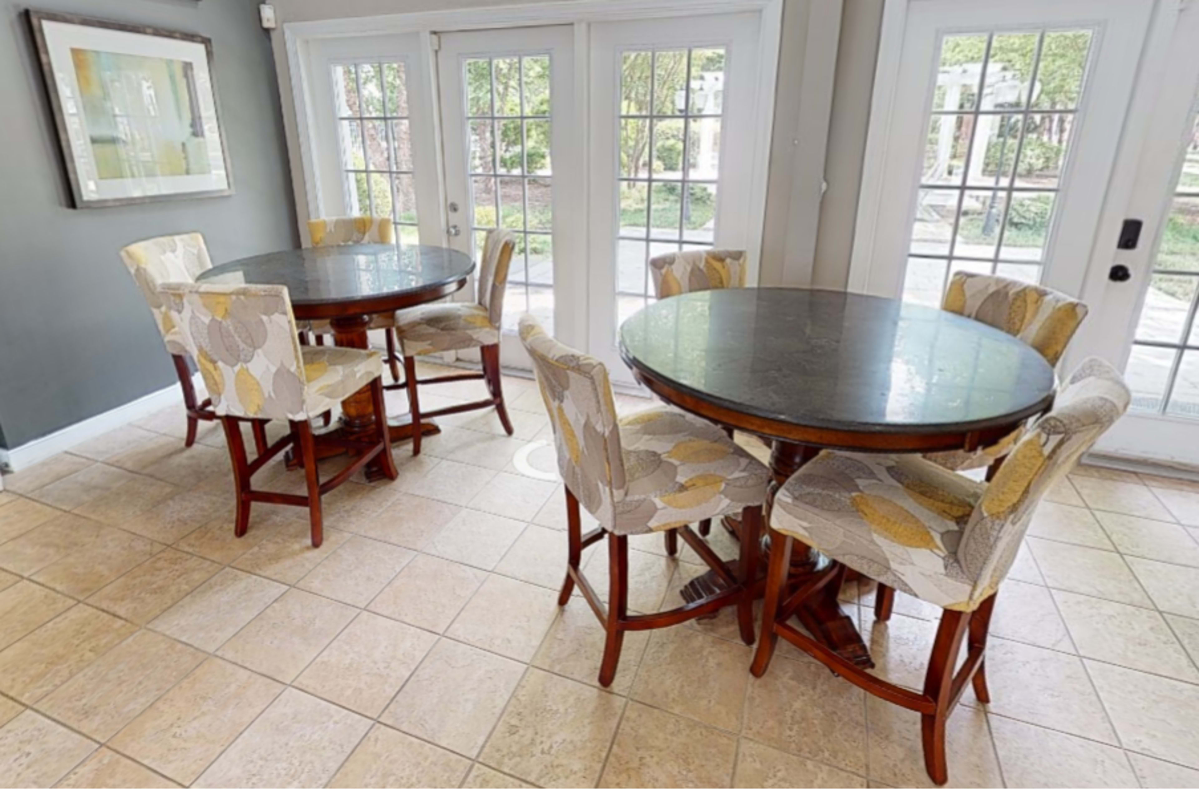 A bright room with two round dining tables, each surrounded by six upholstered chairs, and large windows allowing natural light to enter.