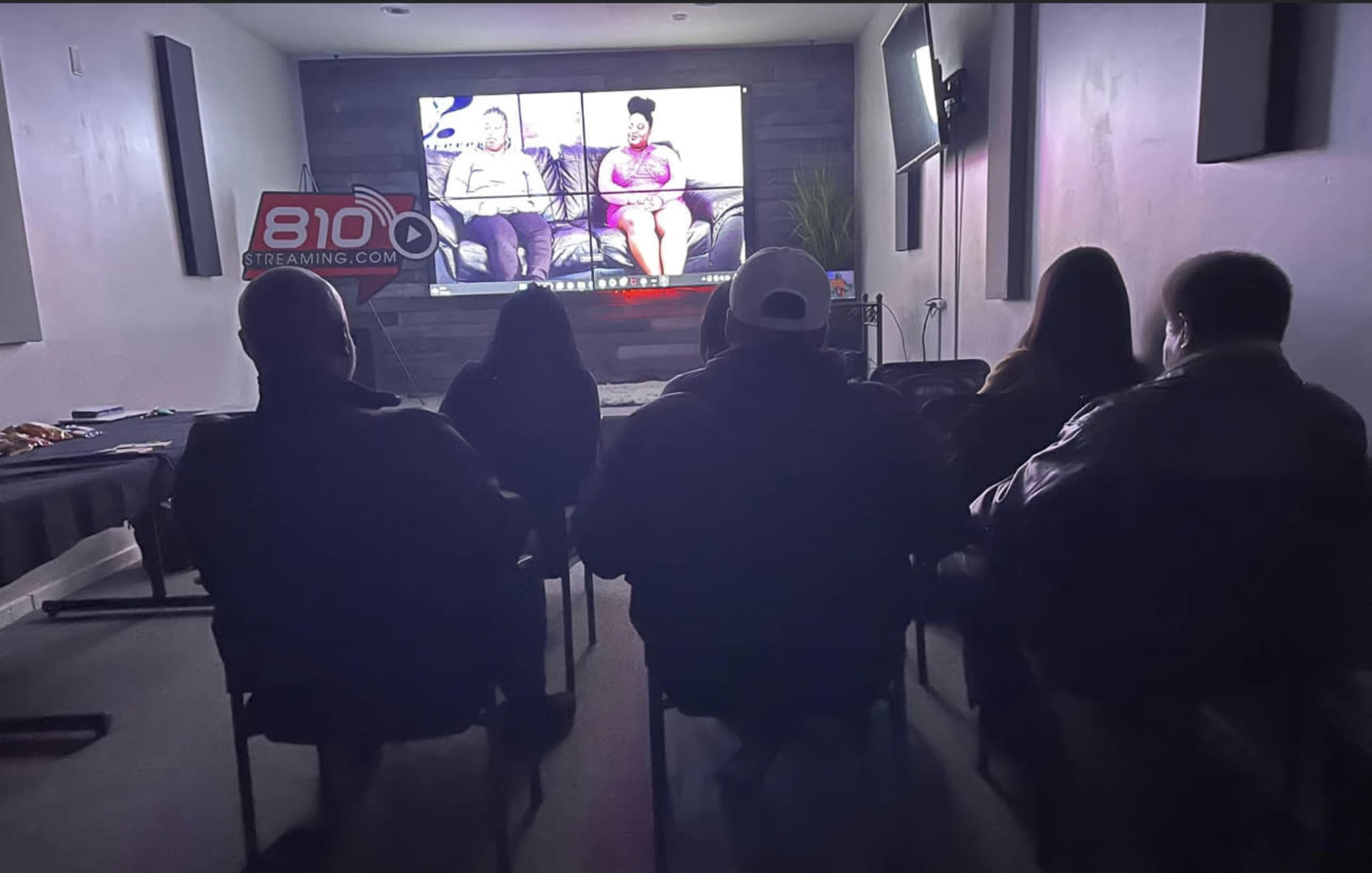 A group of people sits in chairs facing a large screen that displays two individuals on a split screen.