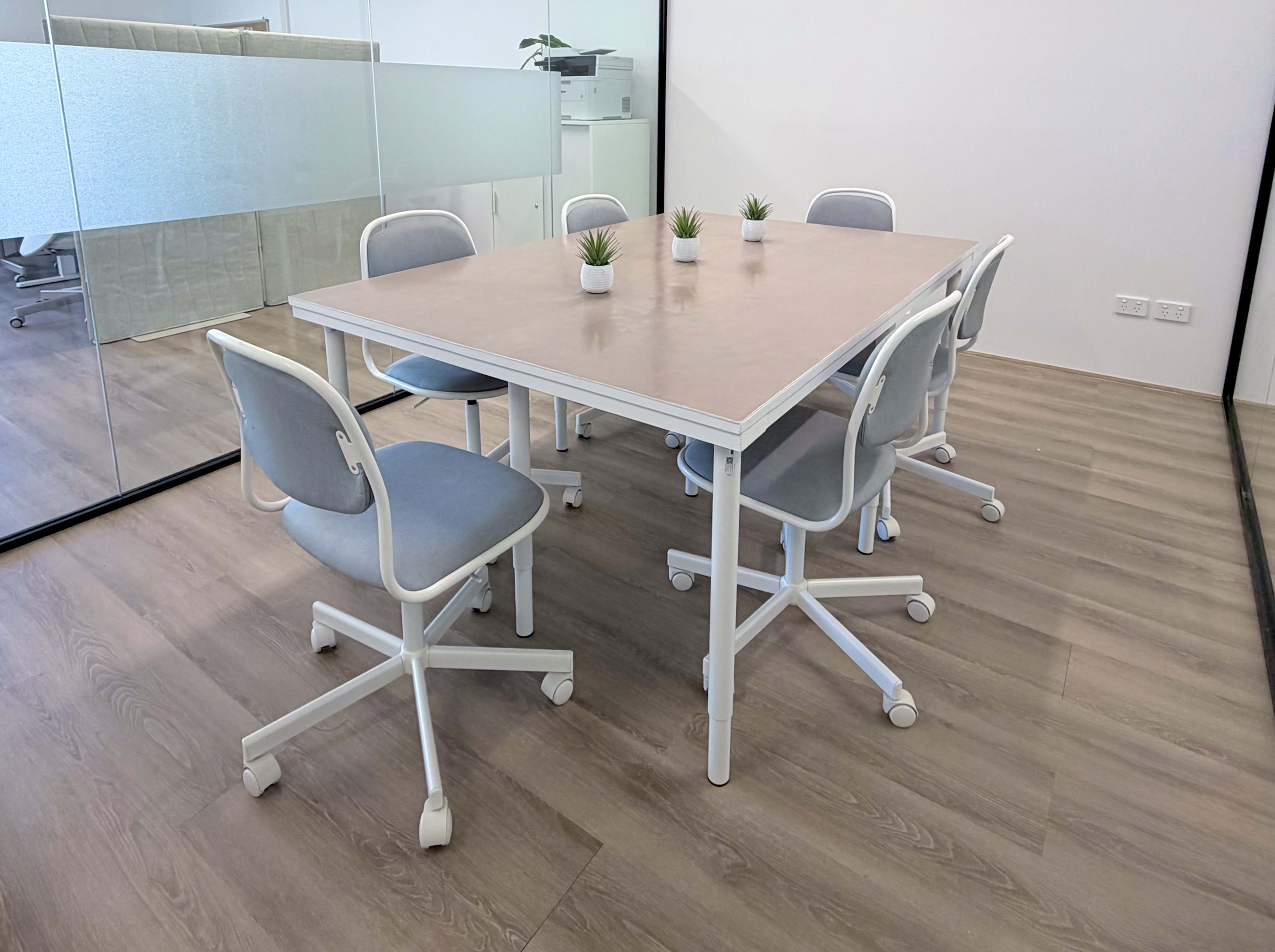 A modern conference room features a light wooden table surrounded by six chair with wheels, each adorned with a small potted plant.