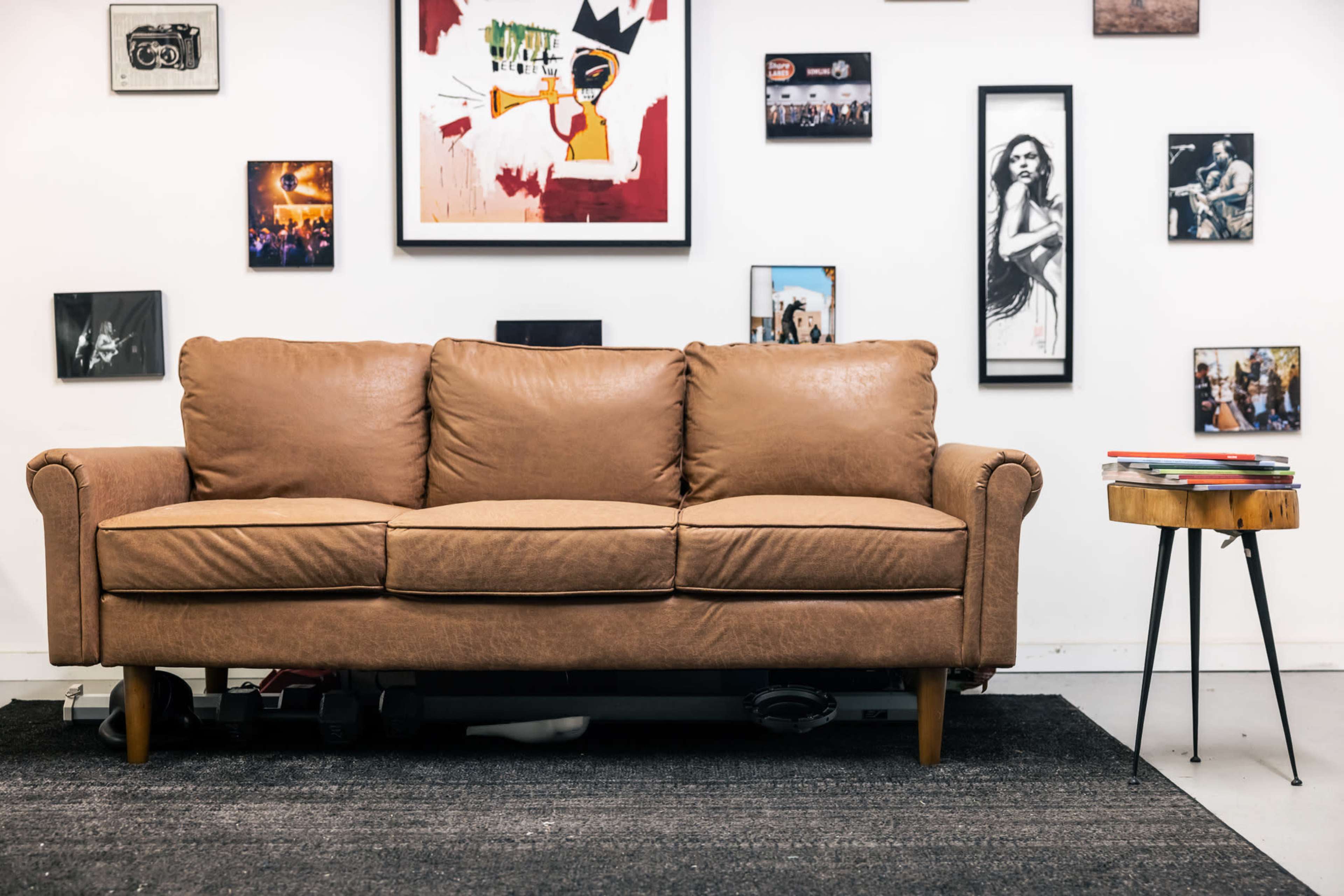 A brown couch is positioned in front of a wall adorned with various framed artworks and photographs, alongside a small side table with books.