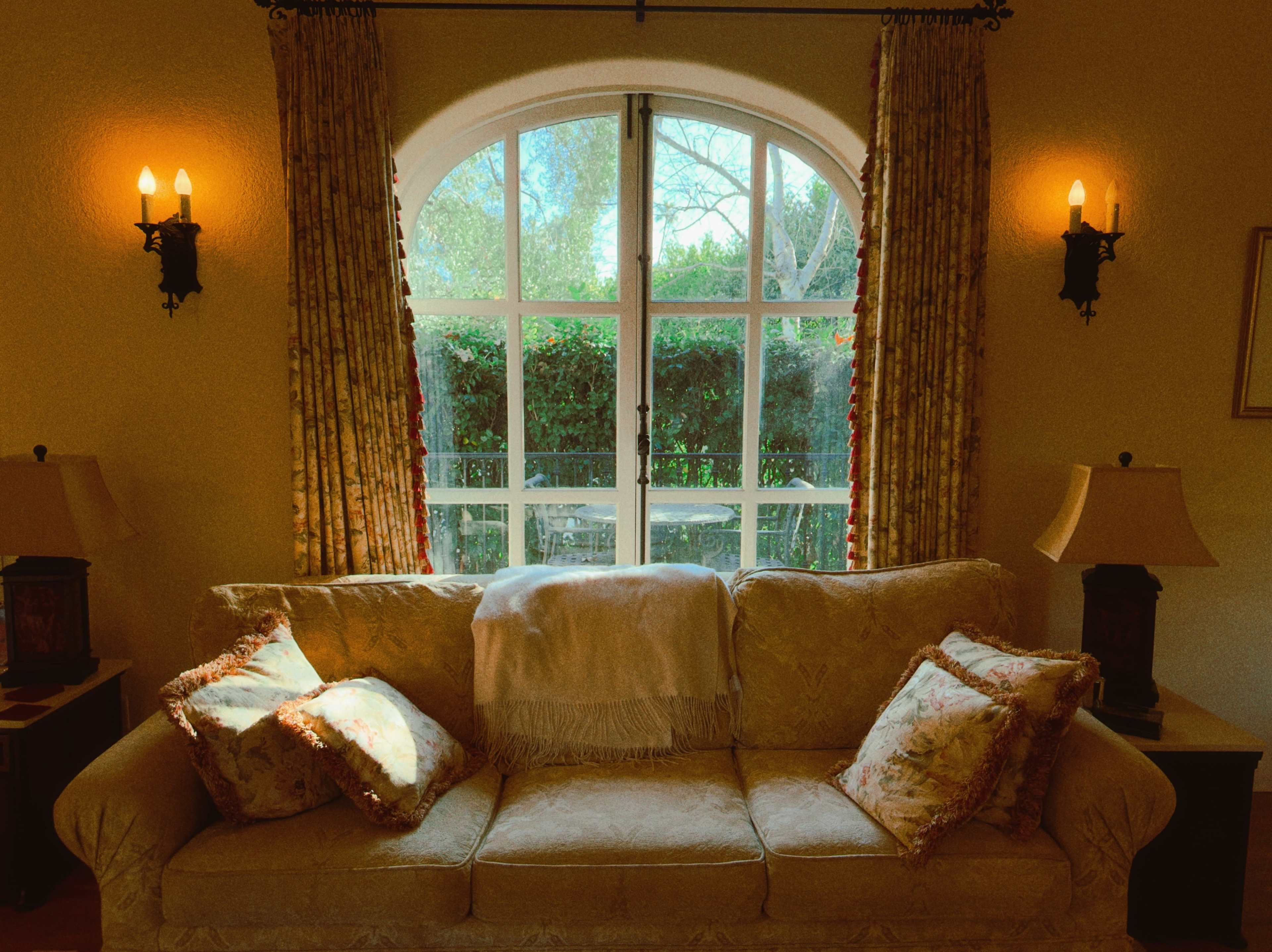 The image shows a cozy living room featuring a light-colored couch with decorative pillows, two lamps on side tables, and a large arched window framed by floral curtains.