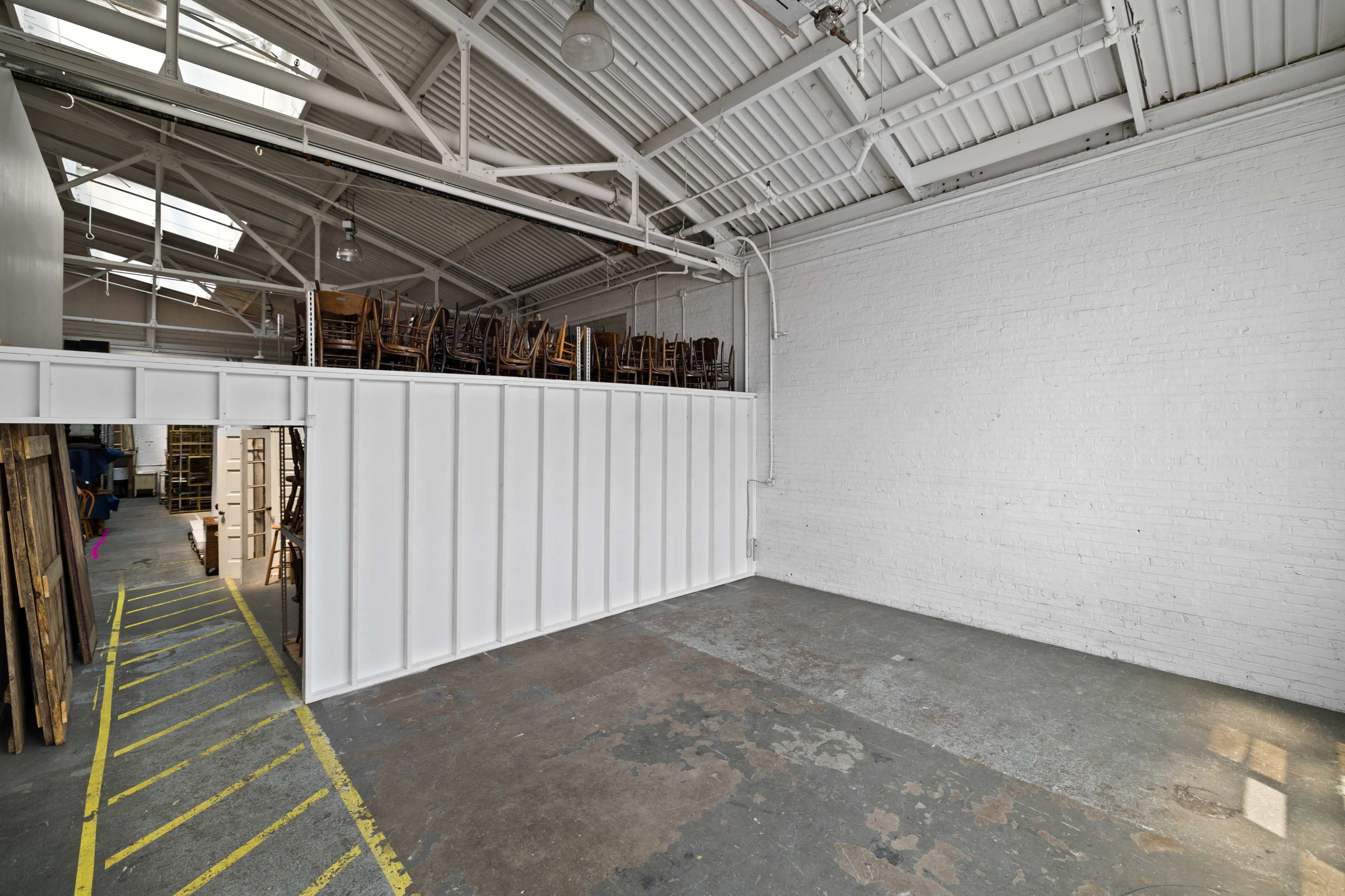 An empty warehouse with a high ceiling, white walls, and a sectioned-off area containing stacked chairs.