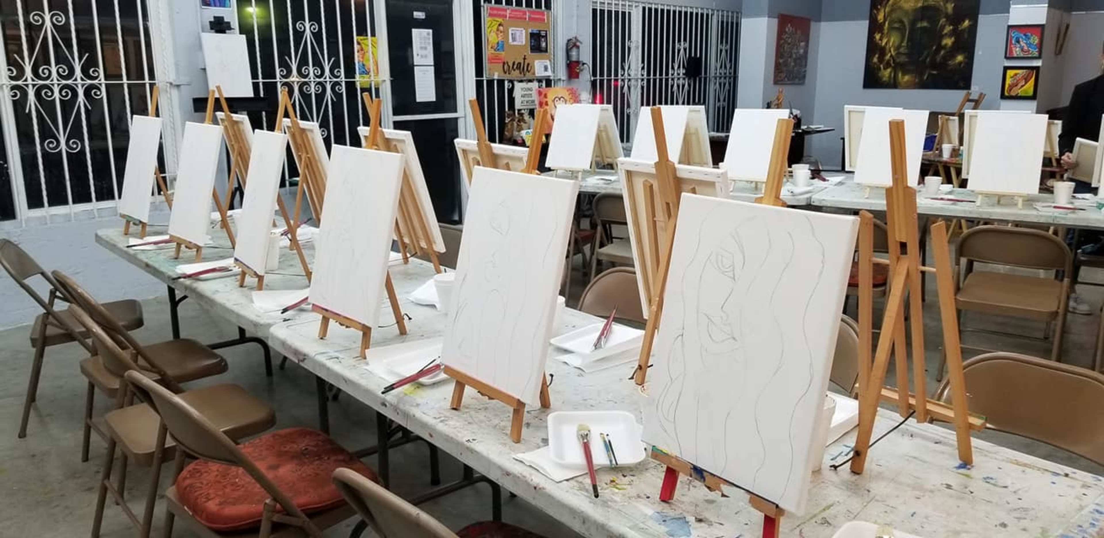 A row of blank canvases on easels set up on tables in an art studio.