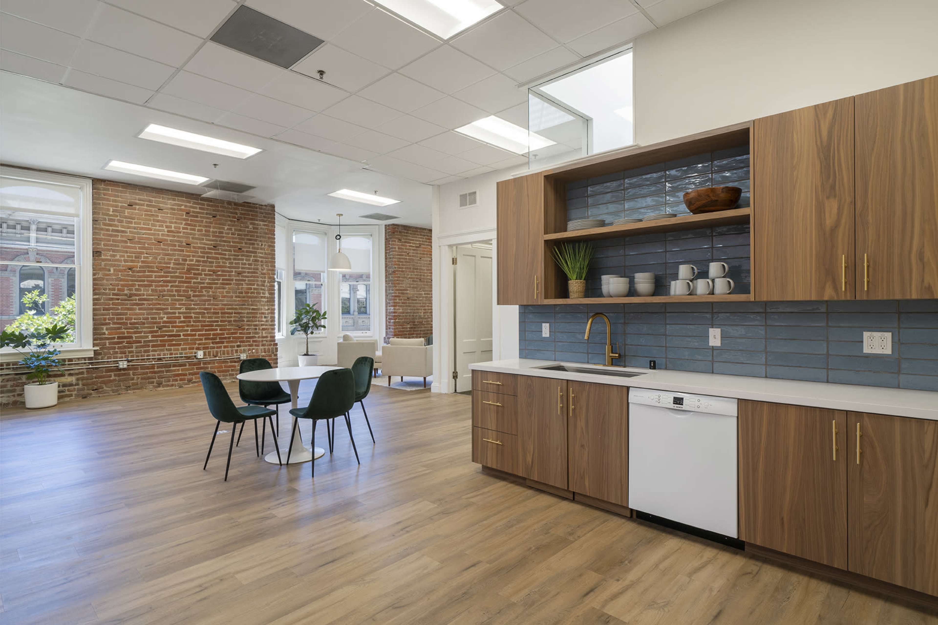 A modern kitchen area with wooden cabinets, a dishwasher, and a dining table with four chairs in a spacious room featuring exposed brick walls and large windows.