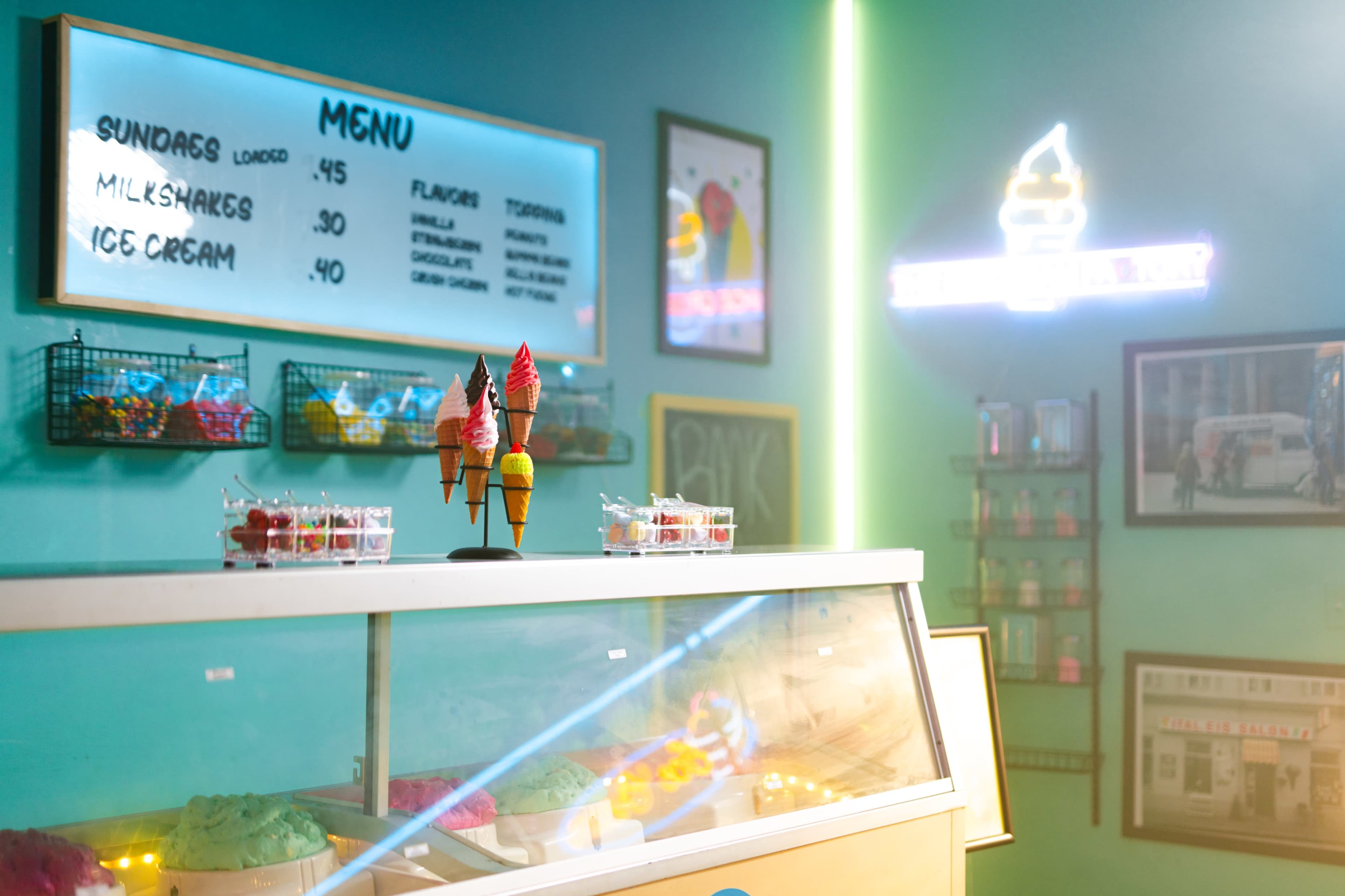 The image shows a brightly colored ice cream shop featuring a display case filled with various ice cream flavors and a menu on the wall listing prices for sundae, milkshakes, and ice cream.