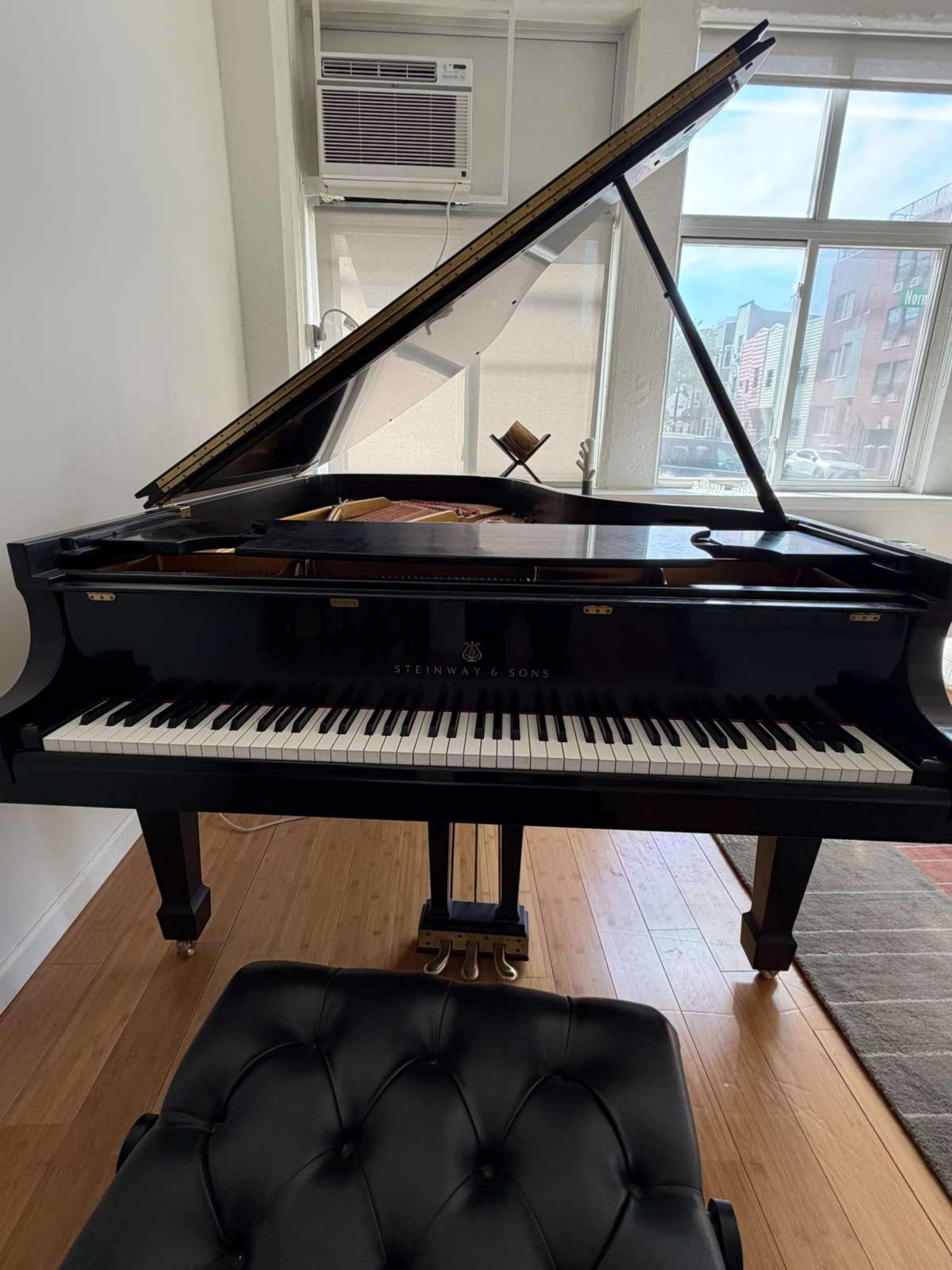 A grand piano is positioned in a well-lit room beside a window, with an air conditioning unit above it and a black cushioned bench nearby.