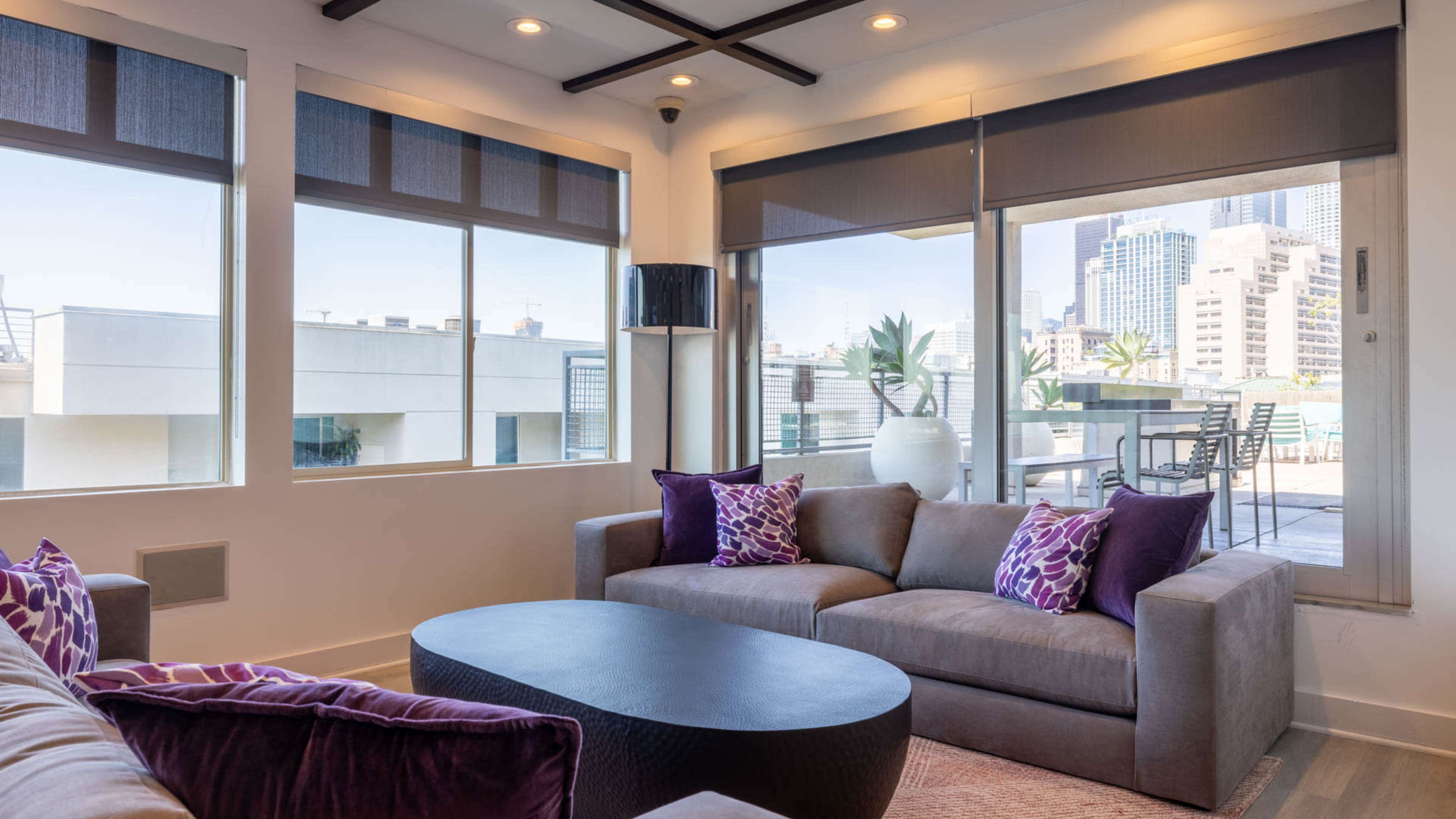A modern living room features a gray sofa with purple pillows, large windows with city views, and a round coffee table.