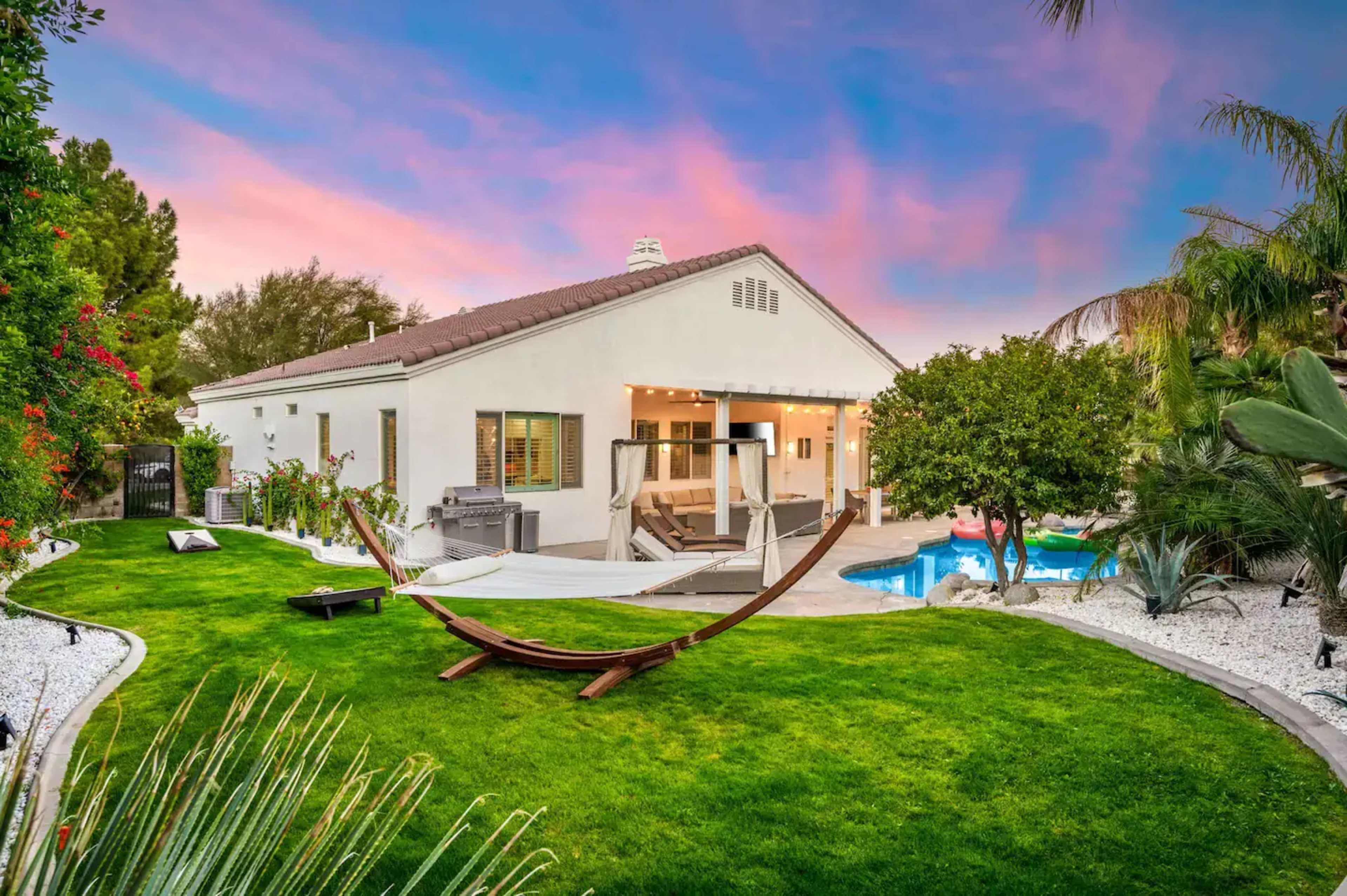 A white house with large windows is set in a landscaped yard featuring a hammock, swimming pool, and various plants against a colorful sunset sky.