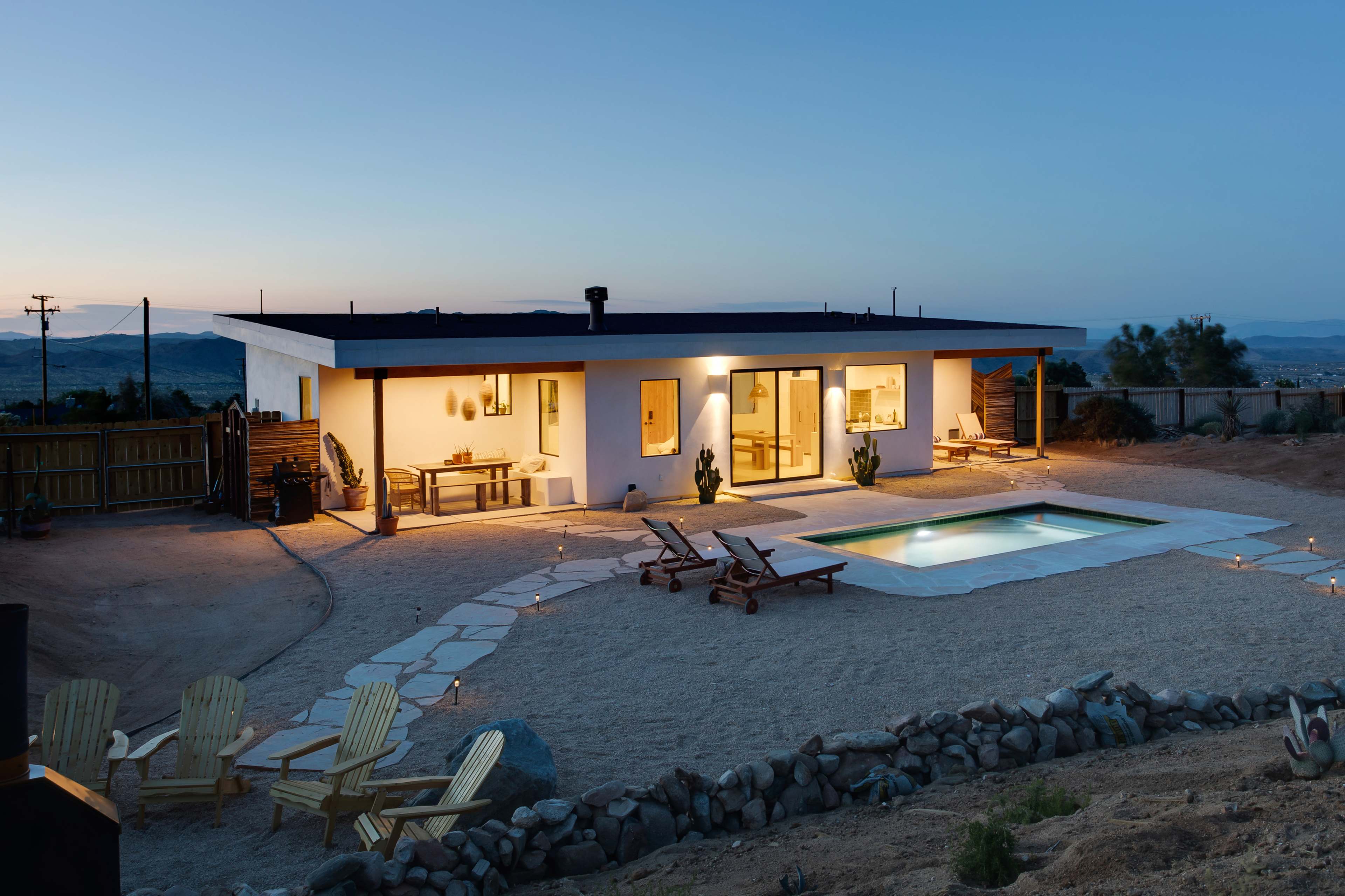 A modern house with a pool is illuminated at dusk, surrounded by a sandy area and desert landscaping.