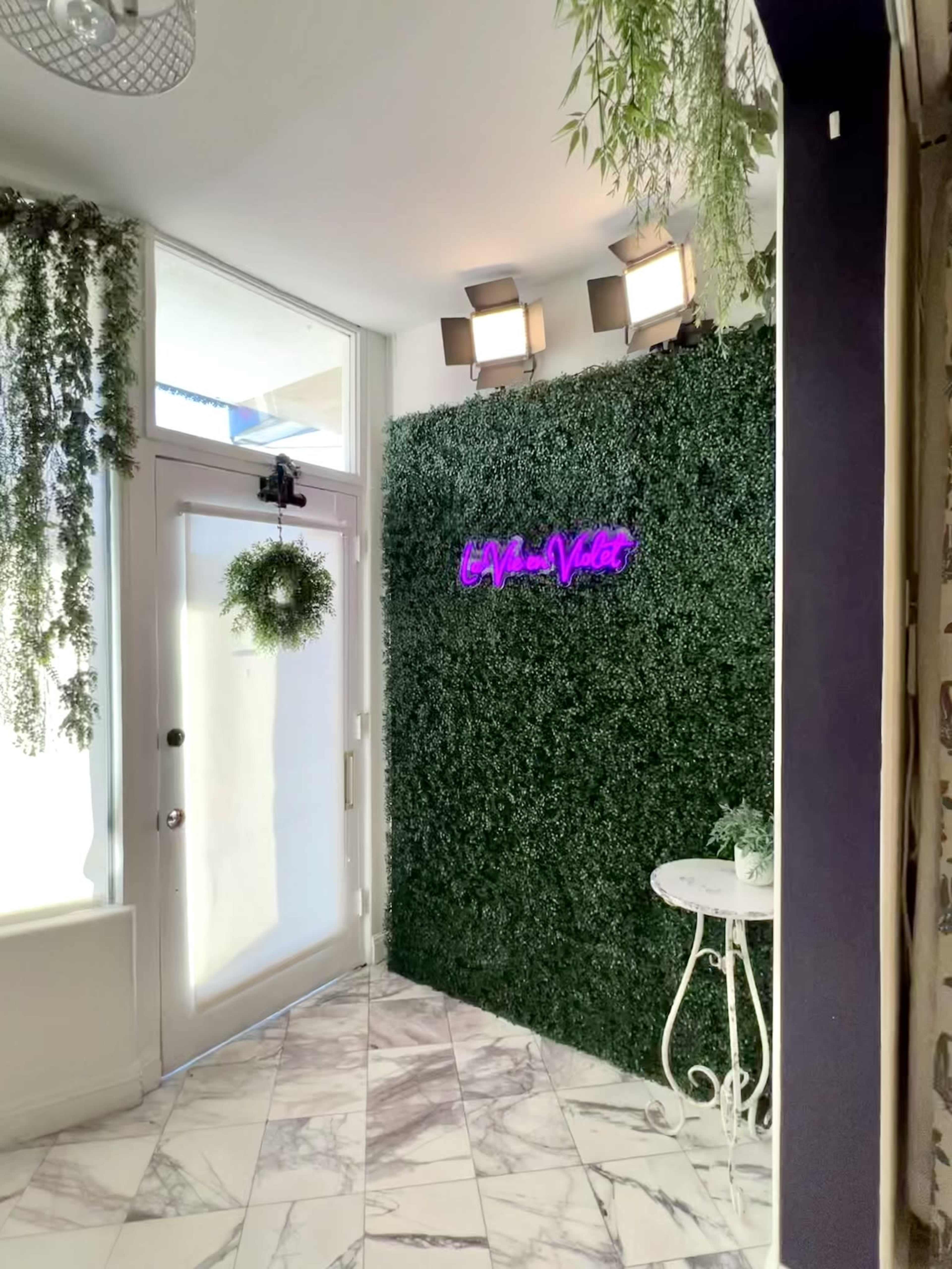 The image shows an entryway with a green wall of artificial foliage, a white door, and a small round table beneath a neon sign.