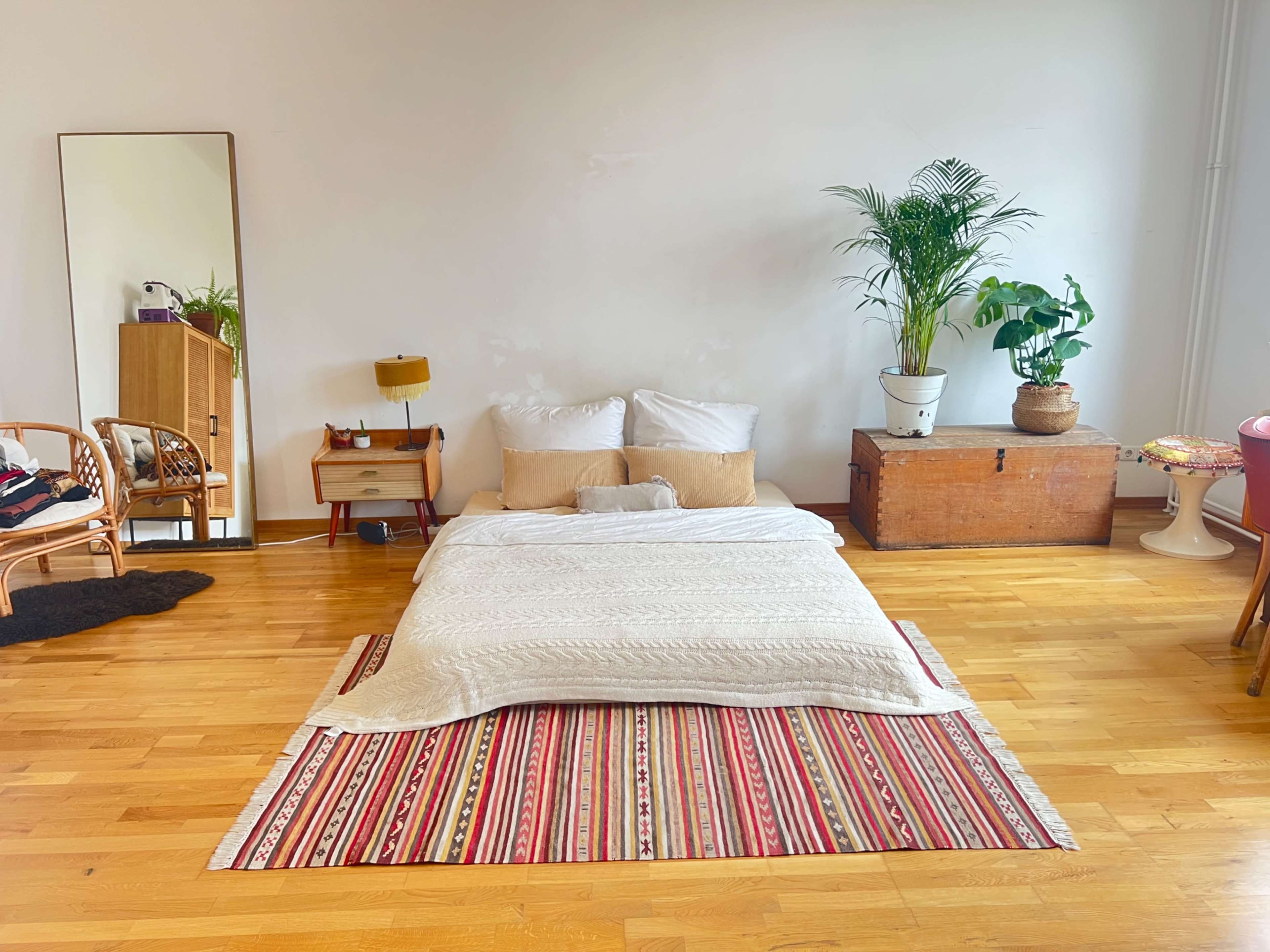 The image shows a simple bedroom with a low bed made of blankets on a colorful rug, surrounded by plants and a mirror.