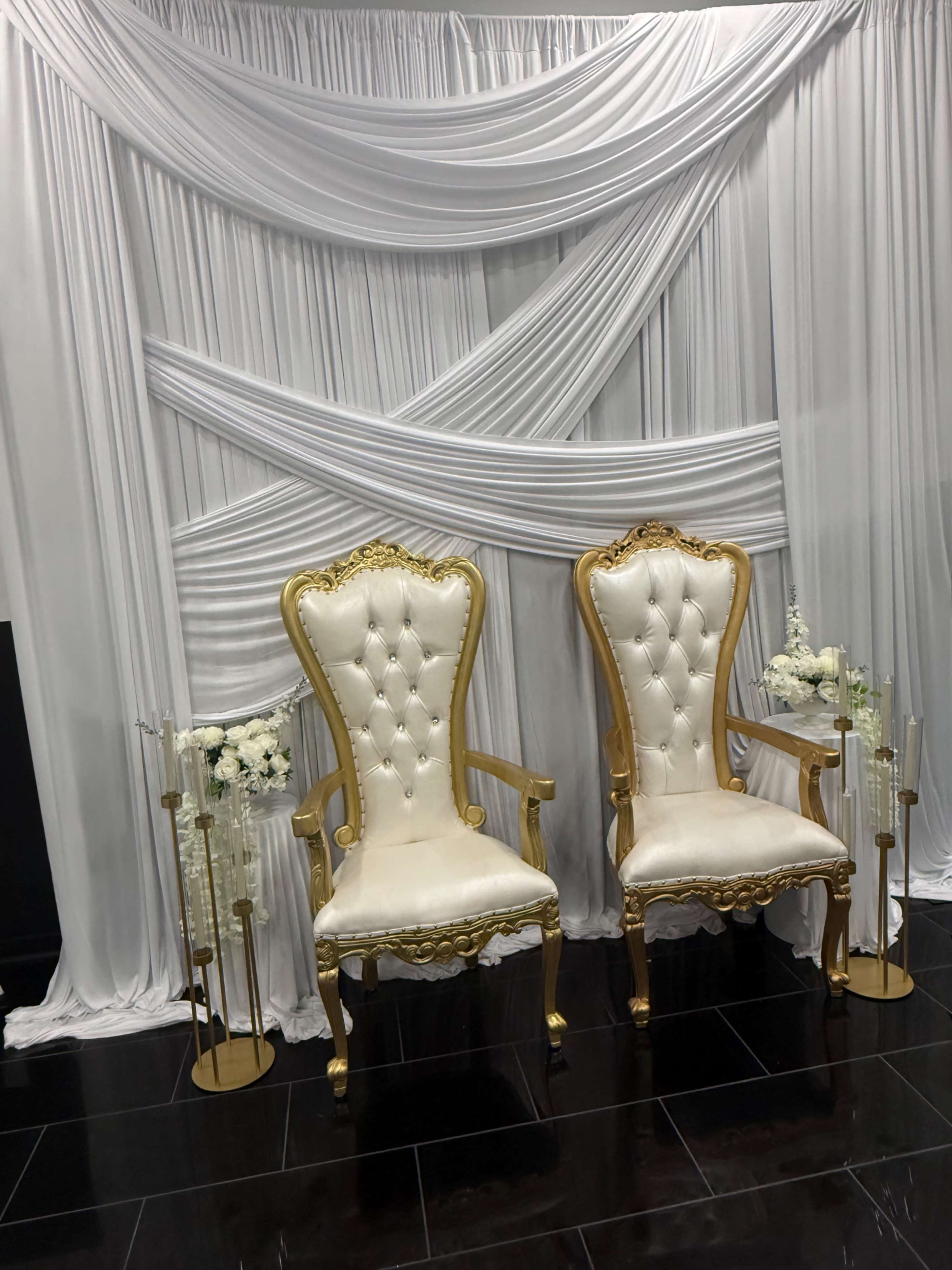 The image shows two ornate golden chairs with white upholstery positioned in front of a backdrop of draped white fabric.