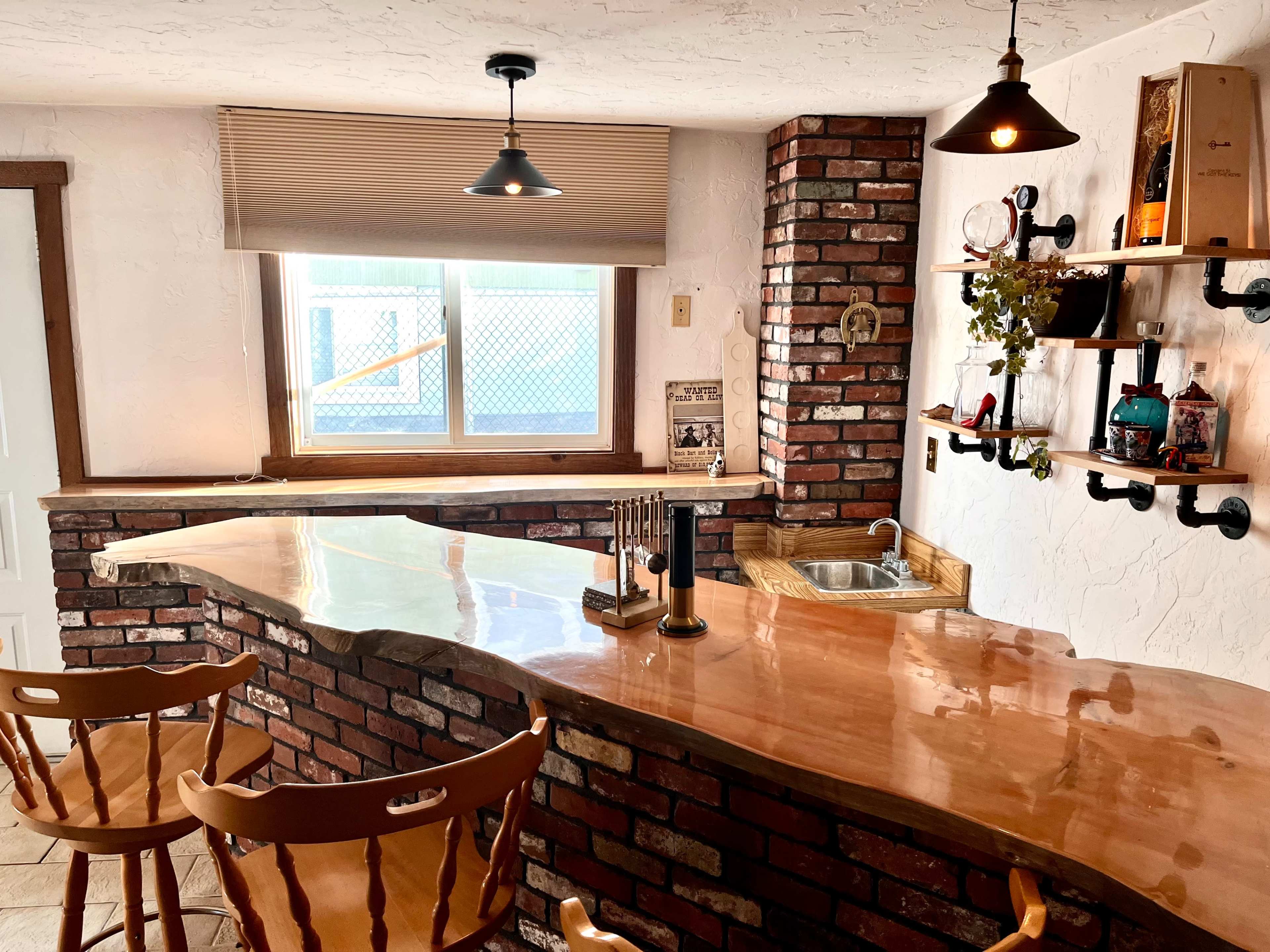 The image shows a small bar area with a curved wooden countertop, brick walls, and two wooden stools, illuminated by hanging light fixtures.