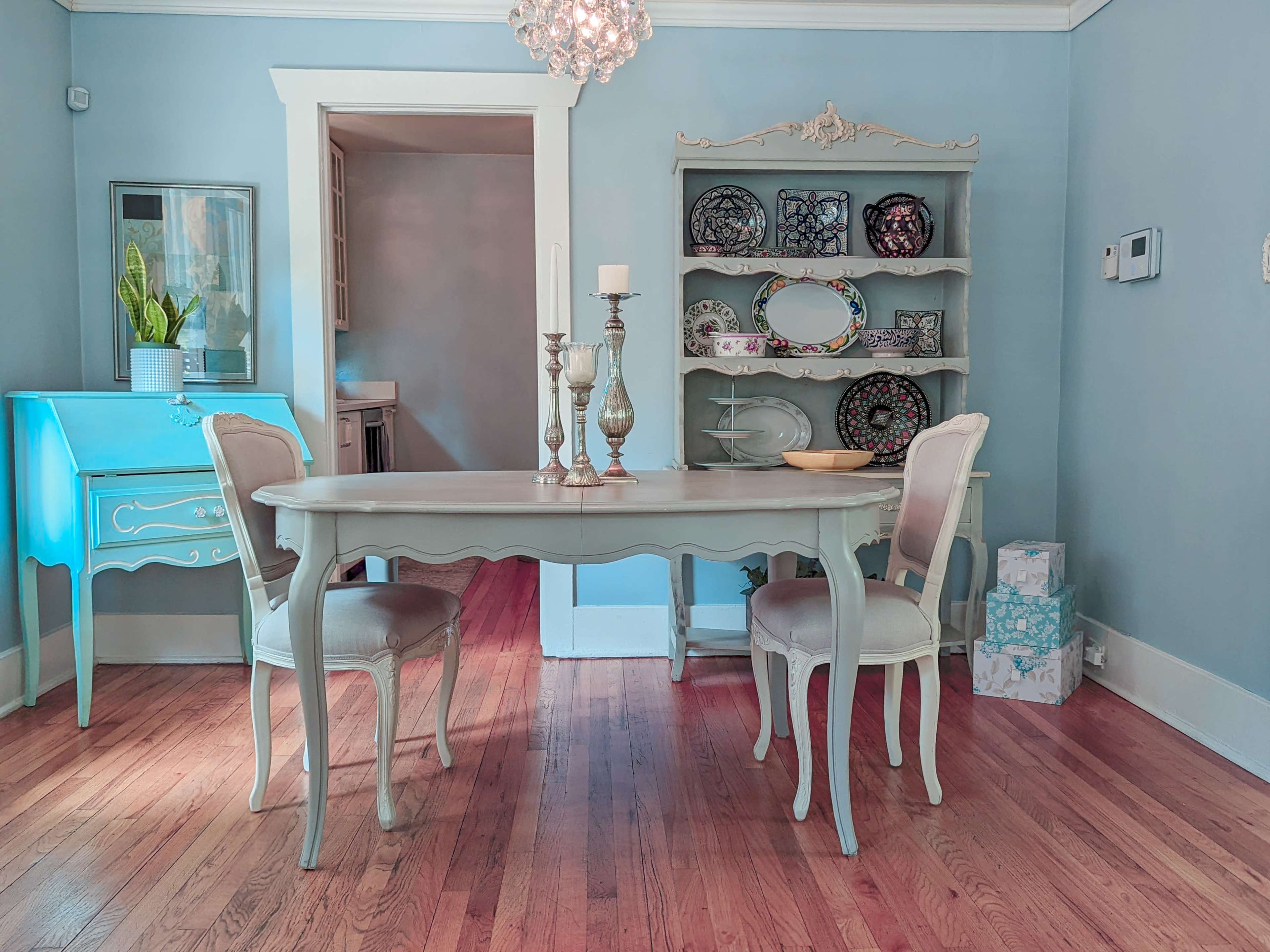 A light blue dining area features a table with two chairs, a decorative shelf filled with plates, and a side cabinet.