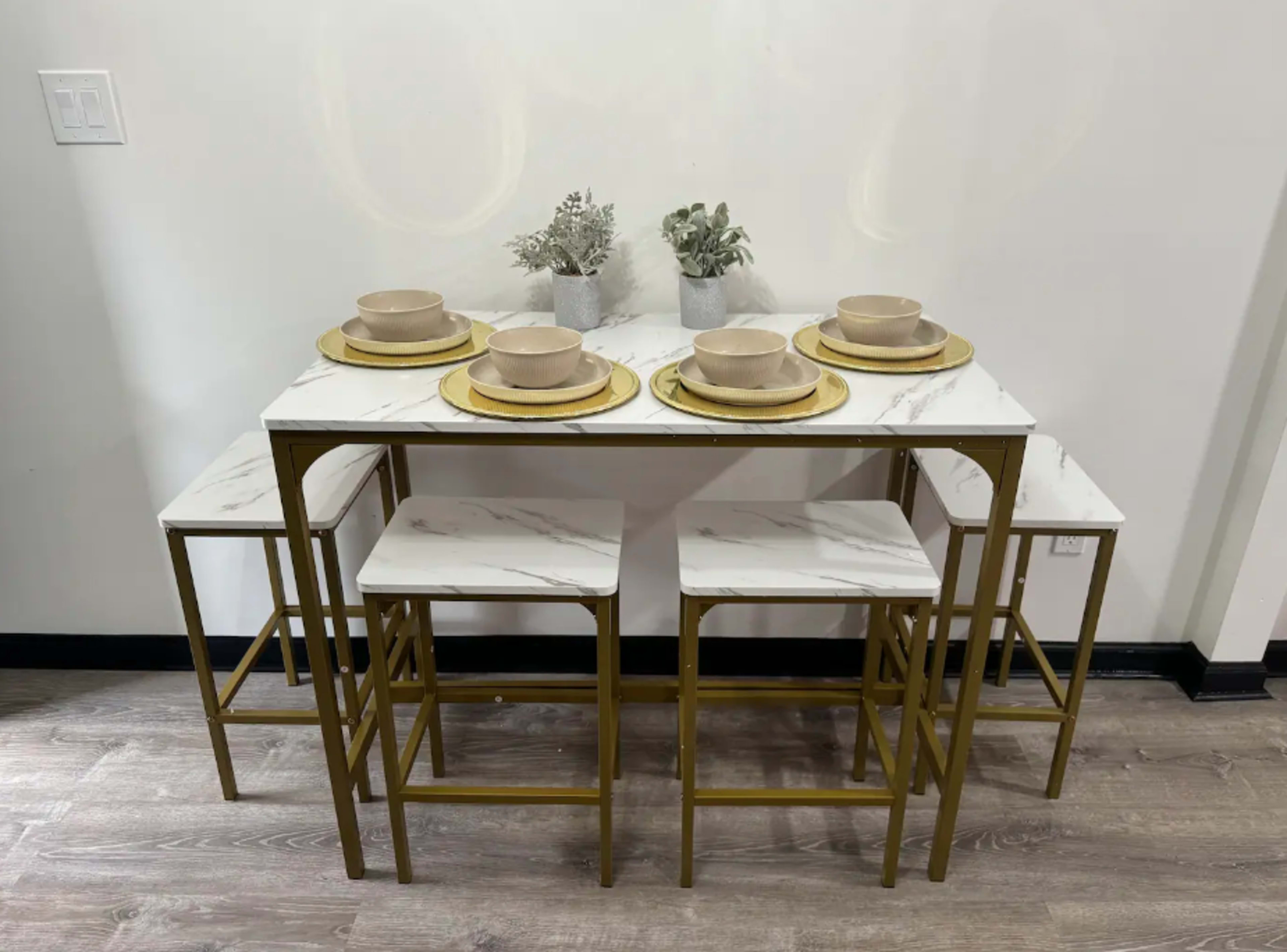 A modern dining table with four gold-framed chairs is set with beige plates and bowls, topped with decorative serveware, against a simple wall.