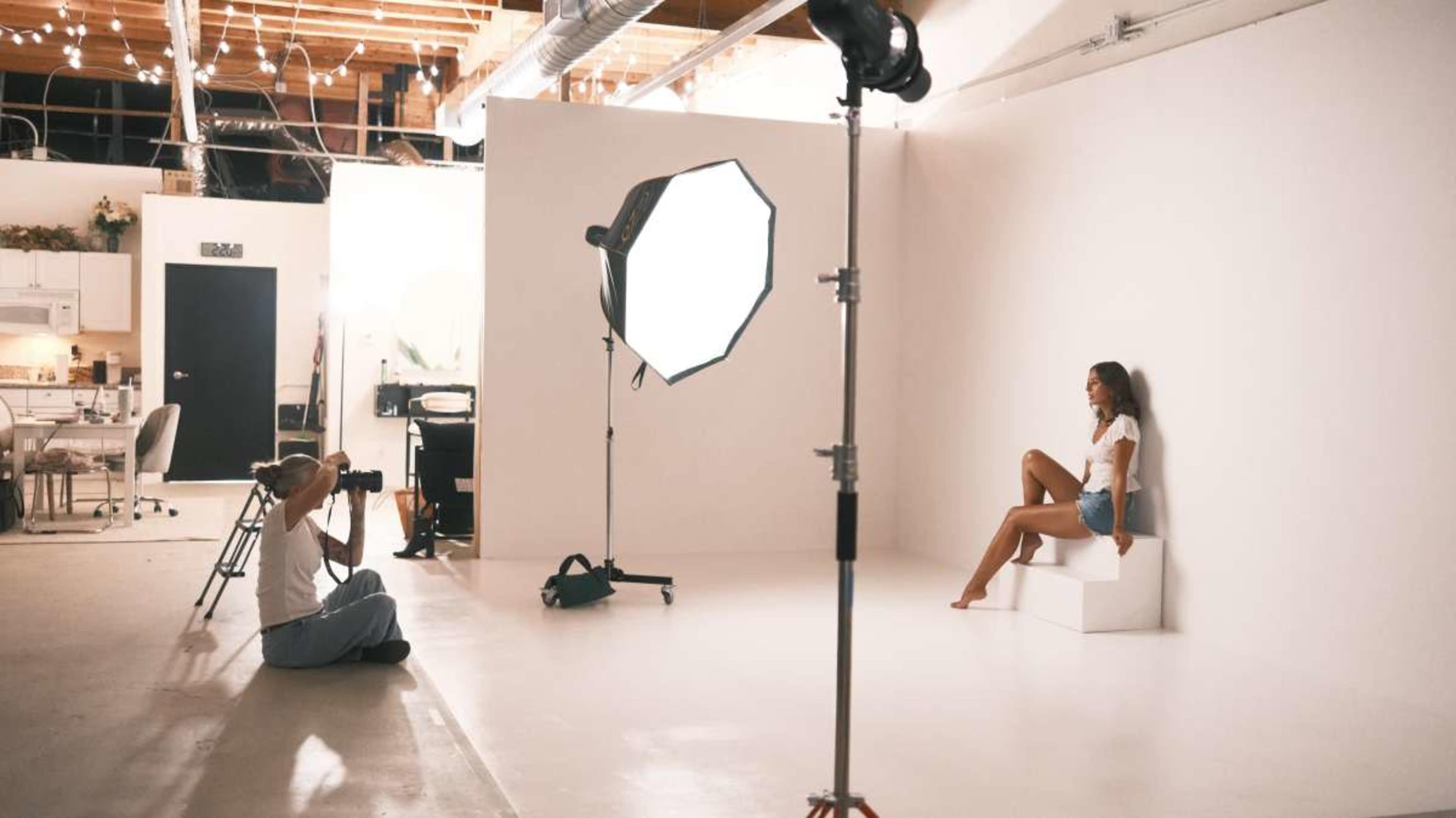 A photographer captures a model posing on a white pedestal in a brightly lit studio with softbox lighting and a simple backdrop.